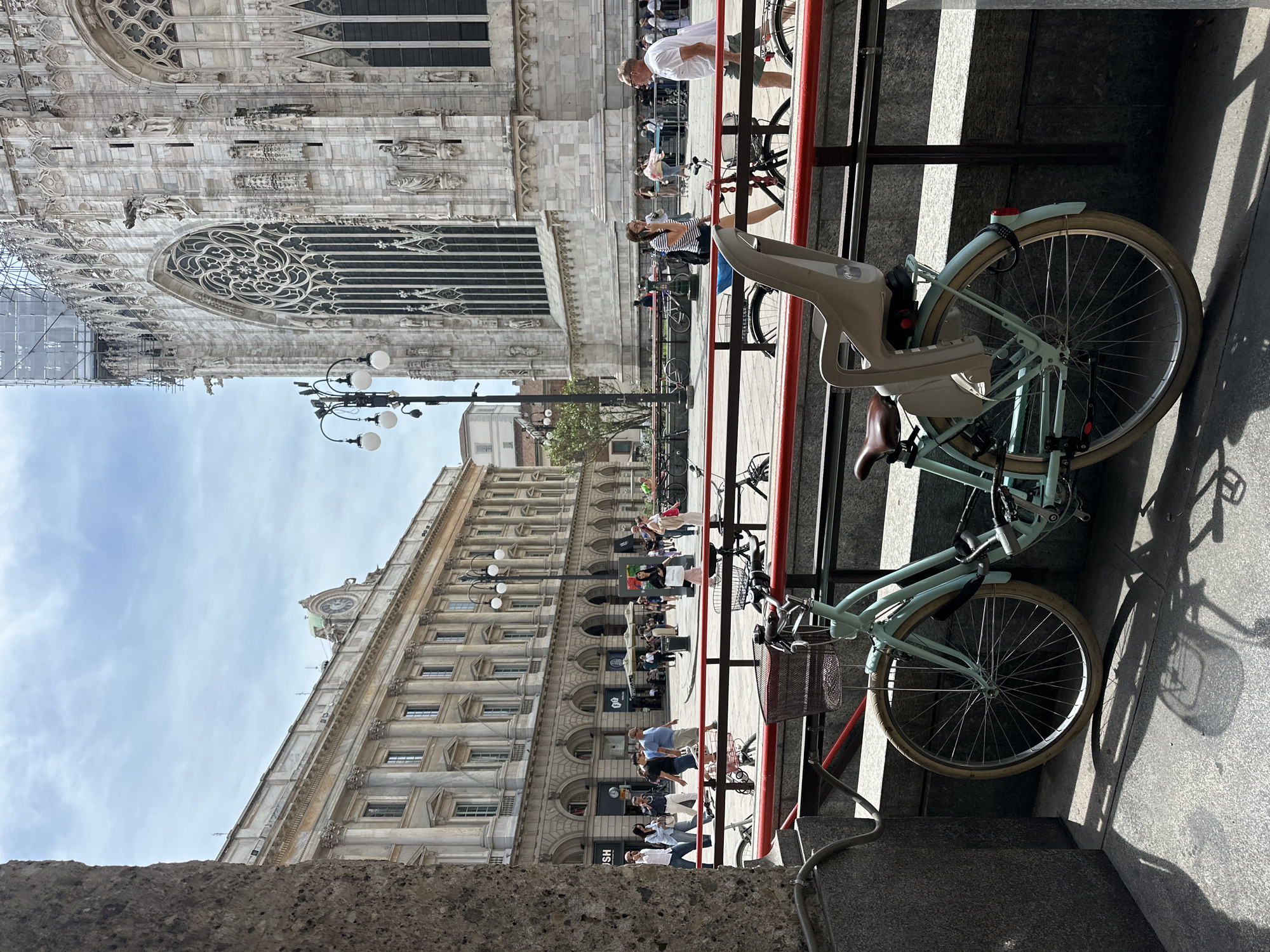 The image captures a scene in Milan, Italy, likely near the Duomo. In the foreground, a light blue bicycle with a child seat is parked, secured with a U-lock. The bicycle is positioned on a paved area, with a red railing behind it. In the background, the facade of a grand, ornate building, characteristic of Milanese architecture and possibly the Duomo itself, is visible. People are walking in the distance, with some seated at outdoor cafes. The sky is partly cloudy. The scene depicts a typical urban environment with historical architecture and pedestrian activity.