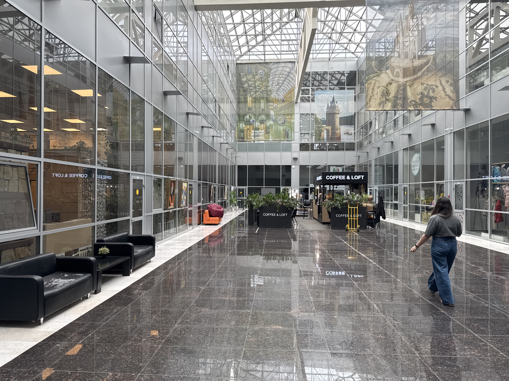 The image shows the interior of a large, modern atrium, possibly a shopping mall or office building, in Moscow, Russia. The space is defined by a glass and steel structure, allowing ample natural light to penetrate through the grid-like ceiling. The walls are lined with large glass panels, some adorned with sizable decorative murals or images.
The floor is made of dark, reflective tiles, and there are seating areas with black leather sofas and small tables. Several businesses, including a "COFFEE & LOFT" establishment, are visible in the background. Plants in large rectangular containers decorate the central walkway, guiding traffic.
A woman is walking through the atrium, facing away from the camera and towards the foreground. She is wearing a gray sweater and blue jeans. The atrium looks fairly empty, with only a few other people visible in the distance.
Based on the overall architectural style and the "PIANO MANIA" sign visible in one of the windows, this could be the Metropolis shopping mall or a similar modern commercial space in Moscow.