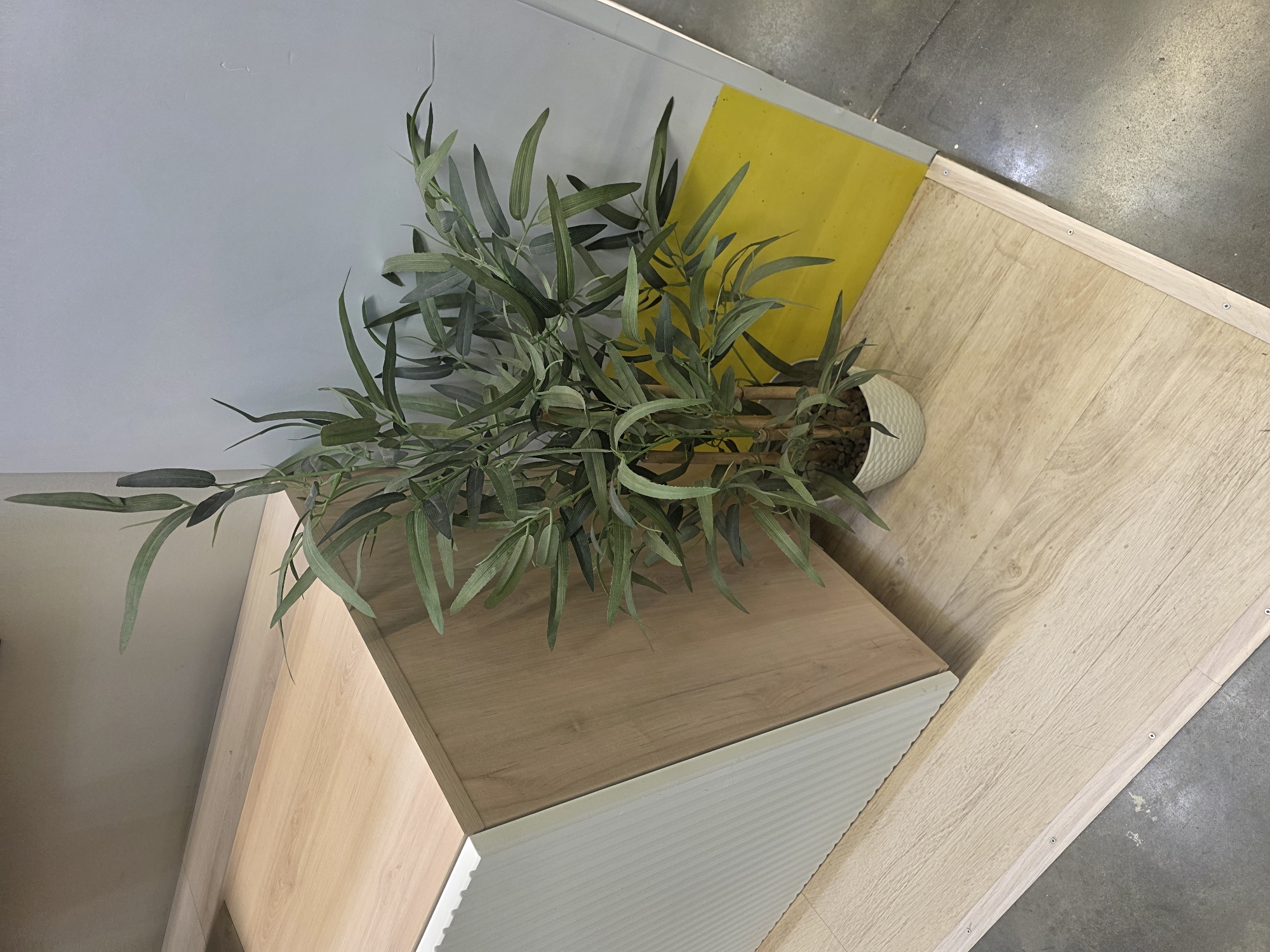 In an indoor setting, a potted artificial bamboo plant stands on a light-colored wooden floor. To the left of the plant, a piece of furniture with a white ribbed side and a light wood-grain front is visible. The furniture has a distinct angled edge where the white and wood-grain sides meet. Behind the plant and furniture, a light gray wall transitions into a bright yellow section. The floor continues towards the foreground, with a visible seam where the wooden flooring meets a concrete surface. The overall scene suggests a commercial or display environment, possibly a showroom or retail space.
