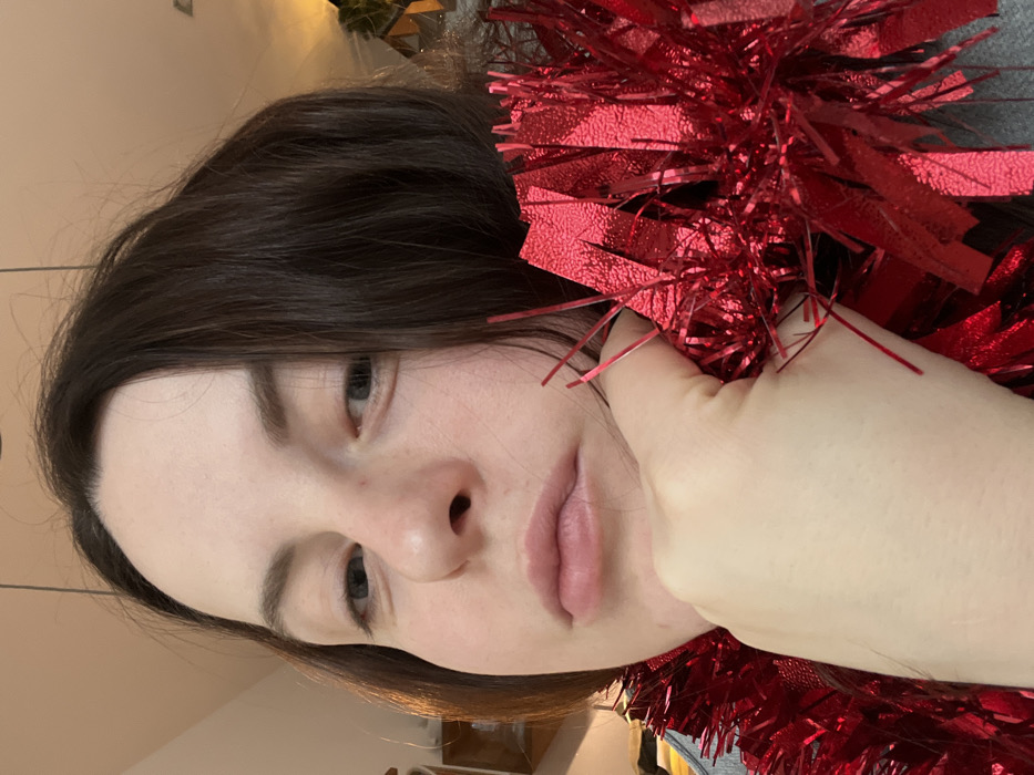 A female individual is depicted in a close-up, head-and-shoulders portrait, in an indoor setting within the city of Callbach, Germany. She has fair skin, dark brown hair, and light-colored eyes, and is looking directly forward with a neutral expression. Her left hand is raised, with her chin resting on the back of her fingers. A large quantity of bright red metallic tinsel or garland is draped around her neck and over her hand. The tinsel is composed of reflective, shimmering red strips. A portion of a grey garment is visible on her left shoulder. The background is blurred, showing indistinct indoor elements and diffuse lighting.