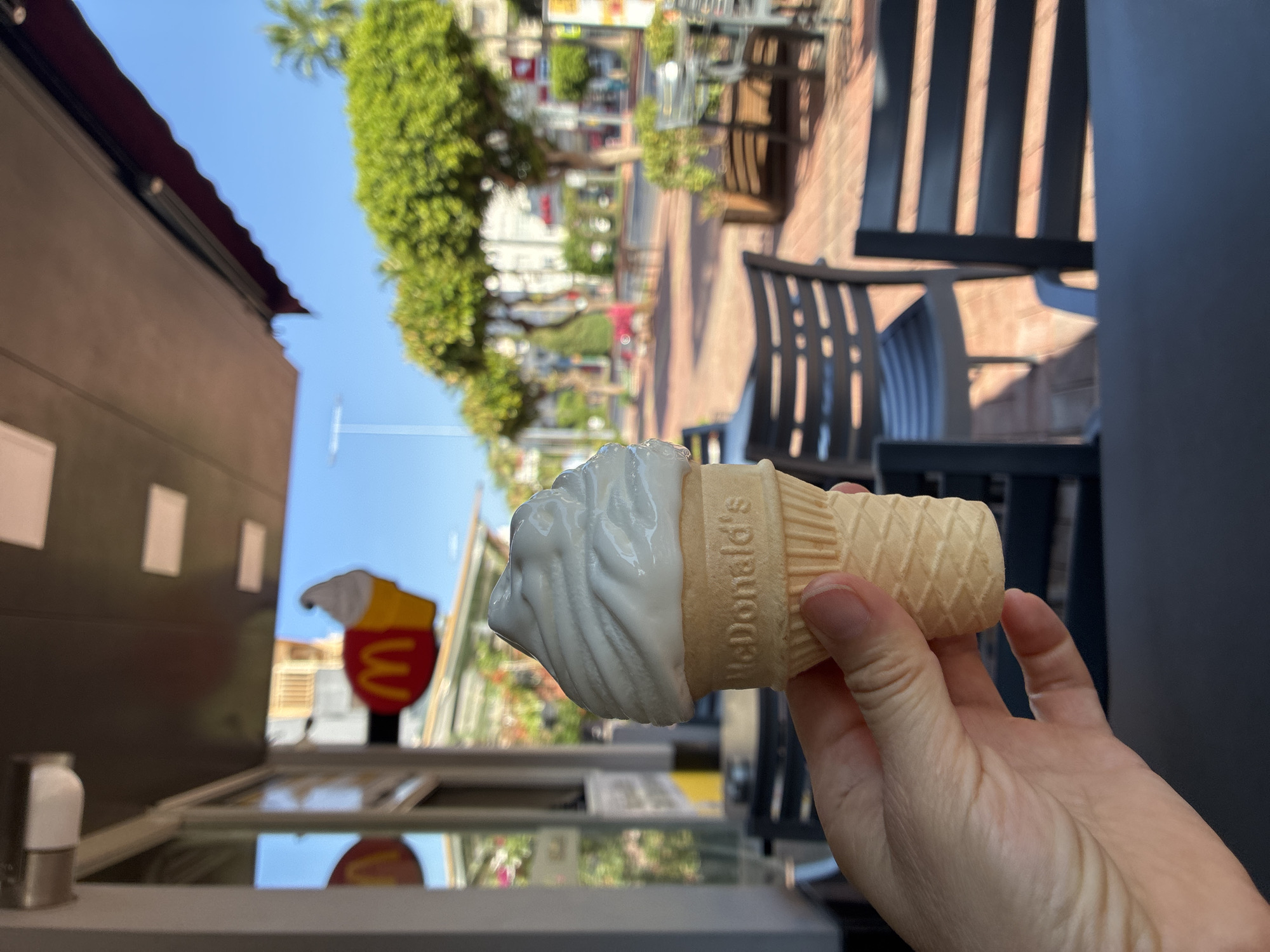 A hand holds a small ice cream cone in front of a McDonald's restaurant in Alanya, Turkey. The ice cream is white and swirled on top, and the cone is light brown with a waffle pattern and the McDonald's logo embossed on it. In the background, the restaurant's exterior is visible, including a sign with the McDonald's logo. Beyond the restaurant, tables and chairs are set up outdoors, and trees line a street under a clear blue sky. The scene appears to be a sunny day.