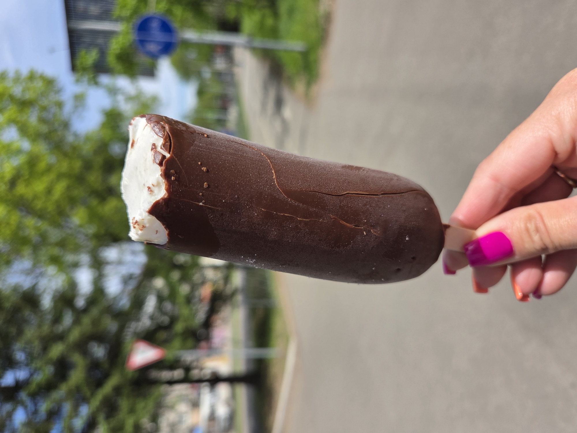 A hand holds a partially eaten chocolate-covered ice cream bar against a blurred background. The ice cream bar is cylindrical with a bite taken out of the top left, revealing white ice cream inside. The chocolate coating has a crack running down its length. The hand holding the ice cream bar has pink and orange nail polish. The background shows a blurred street scene with trees, signs, and a road. The image does not contain any information to verify a location in Birobidzhan, Russia.