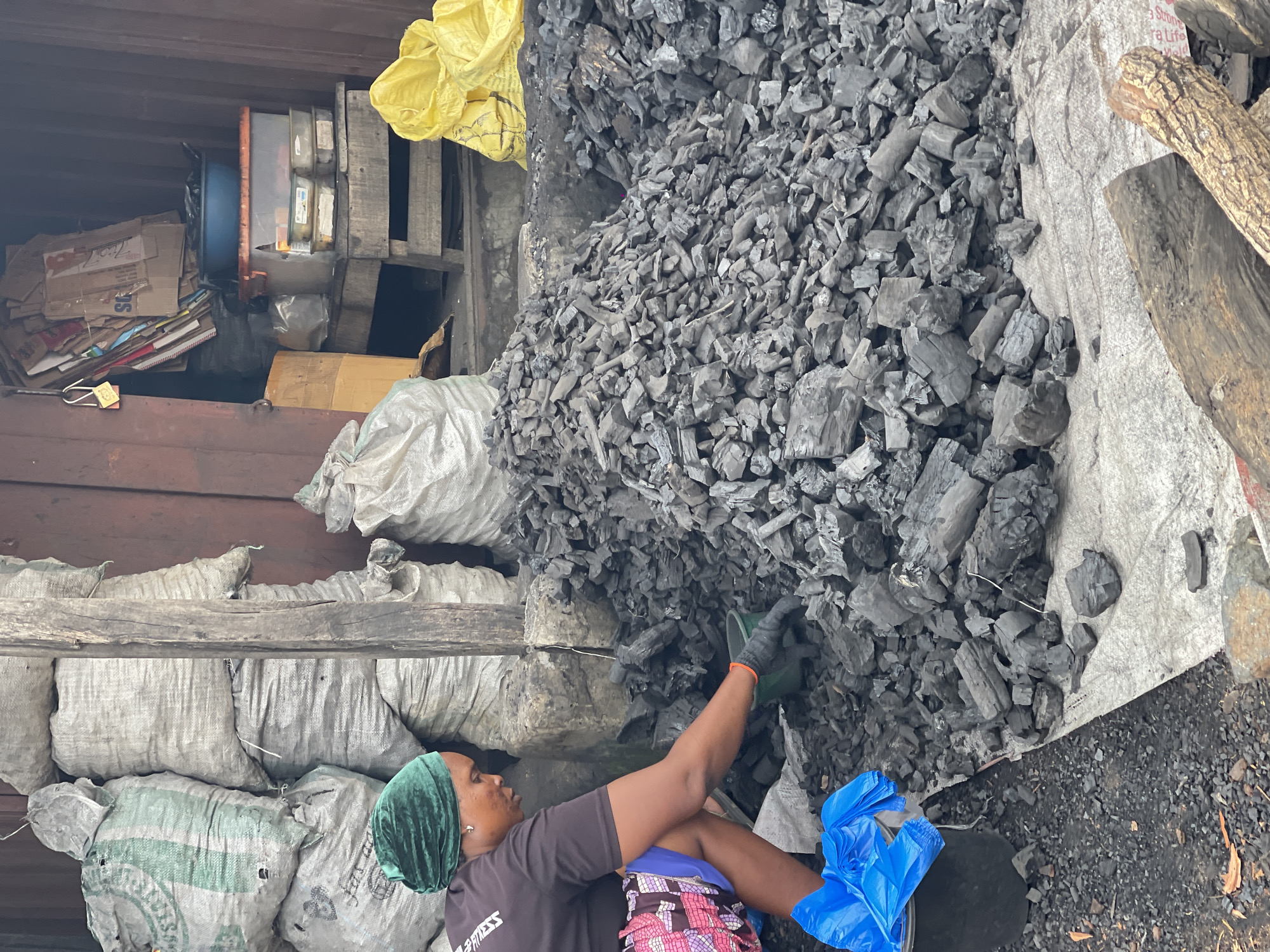 A woman wearing a green headscarf, a dark t-shirt, and a multicolored skirt is crouched on the ground, sorting through a large pile of charcoal. She is wearing black gloves and appears to be placing charcoal into a small green bucket. To her left is a black bucket filled with blue plastic bags. Behind her, stacks of burlap sacks are piled high, some appearing to be filled with charcoal or other materials. In the background, a stall or shop is visible, with shelves holding various items, including containers and cardboard boxes. The ground is covered with charcoal dust and some scattered pieces of wood. The scene appears to be an outdoor market or storage area, likely in Abuja, Nigeria, given the context.