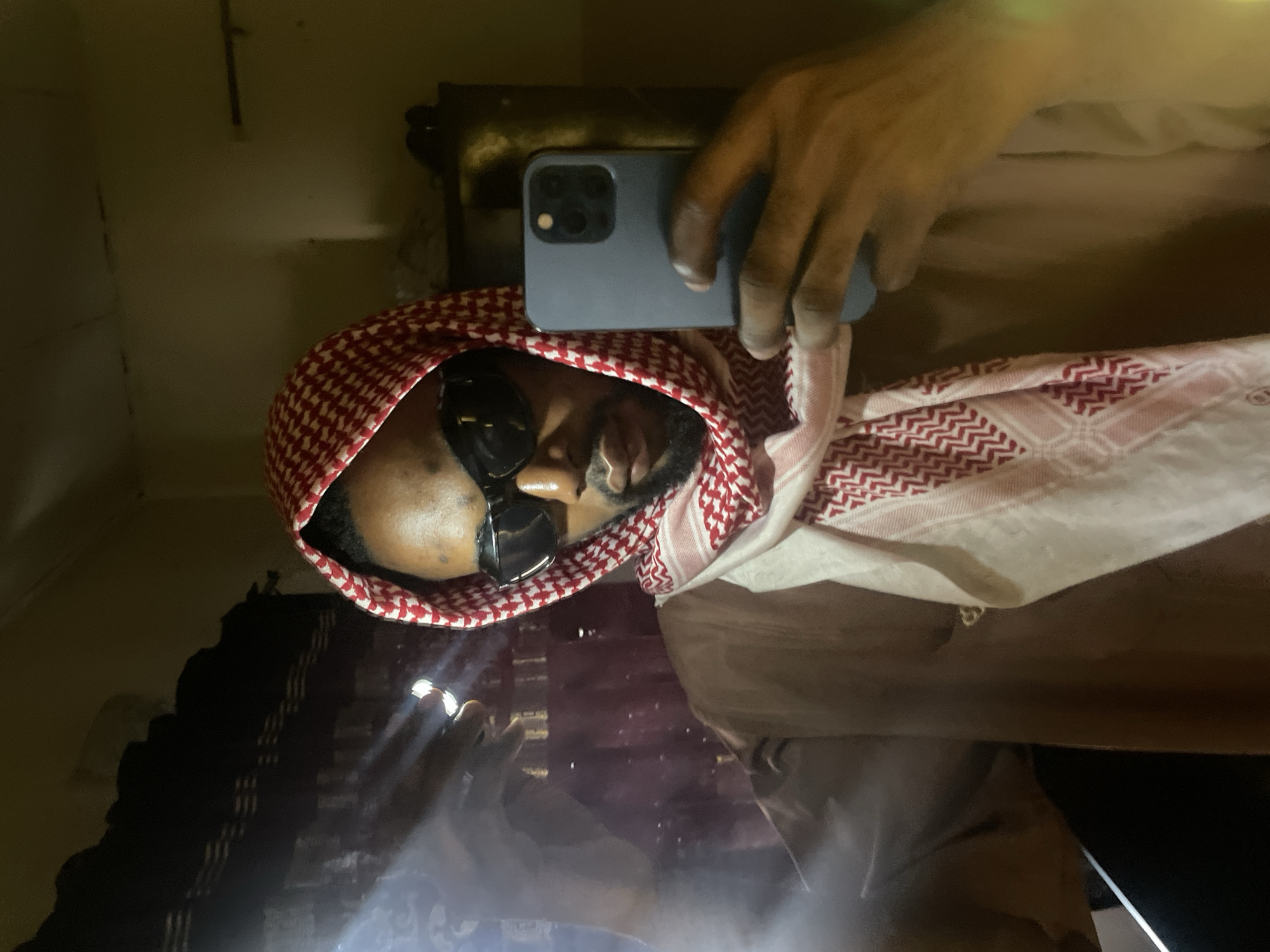 A man with dark skin and facial hair is wearing sunglasses and a red and white checkered headscarf. He is holding a blue smartphone in front of his face, seemingly taking a selfie. The background is dark and indistinct, but there appears to be a reflective surface below him. The setting is not clearly identifiable as Lokoja, Nigeria, without further context.