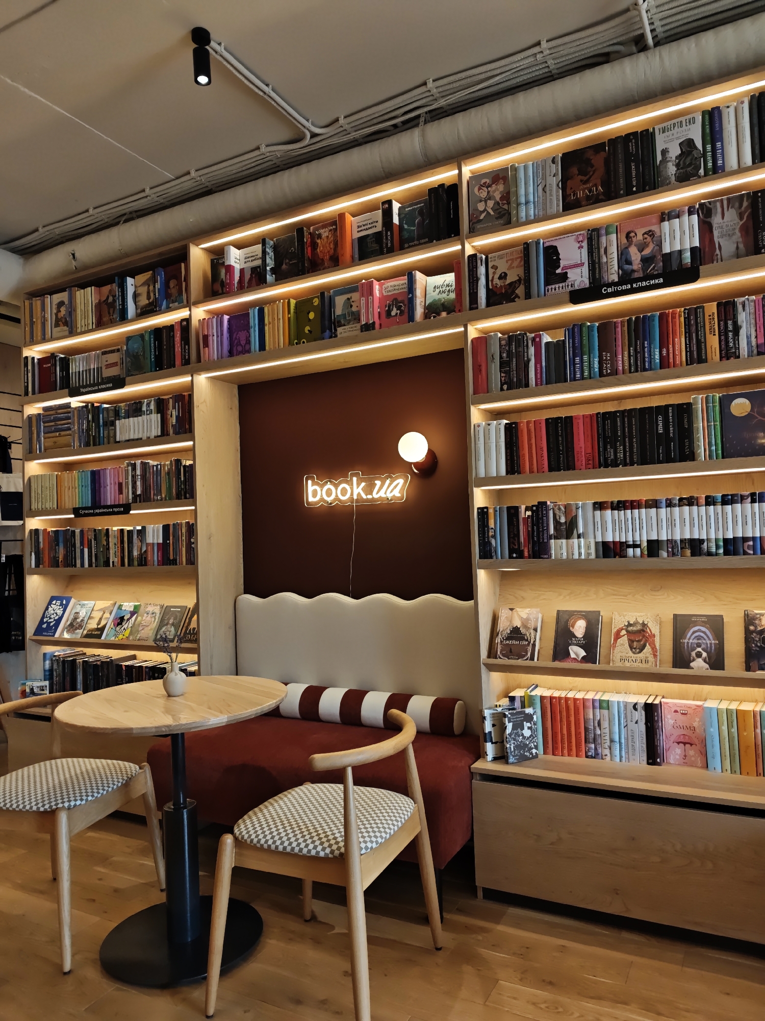 The image shows a cozy corner of a bookstore with wooden bookshelves filled with books. A neon sign that says "book.ua" is mounted on a dark brown wall. In front of the wall, there is a red sofa with white and red striped cushions. A round wooden table with two chairs in front of it is placed in the foreground. The chairs have a black and white checkered pattern on their seats. The overall ambiance suggests a relaxed and inviting atmosphere, possibly a cafe area within a bookstore. The presence of the "book.ua" sign strongly indicates the location is a branch of this bookstore chain, which operates in Kyiv, Ukraine.