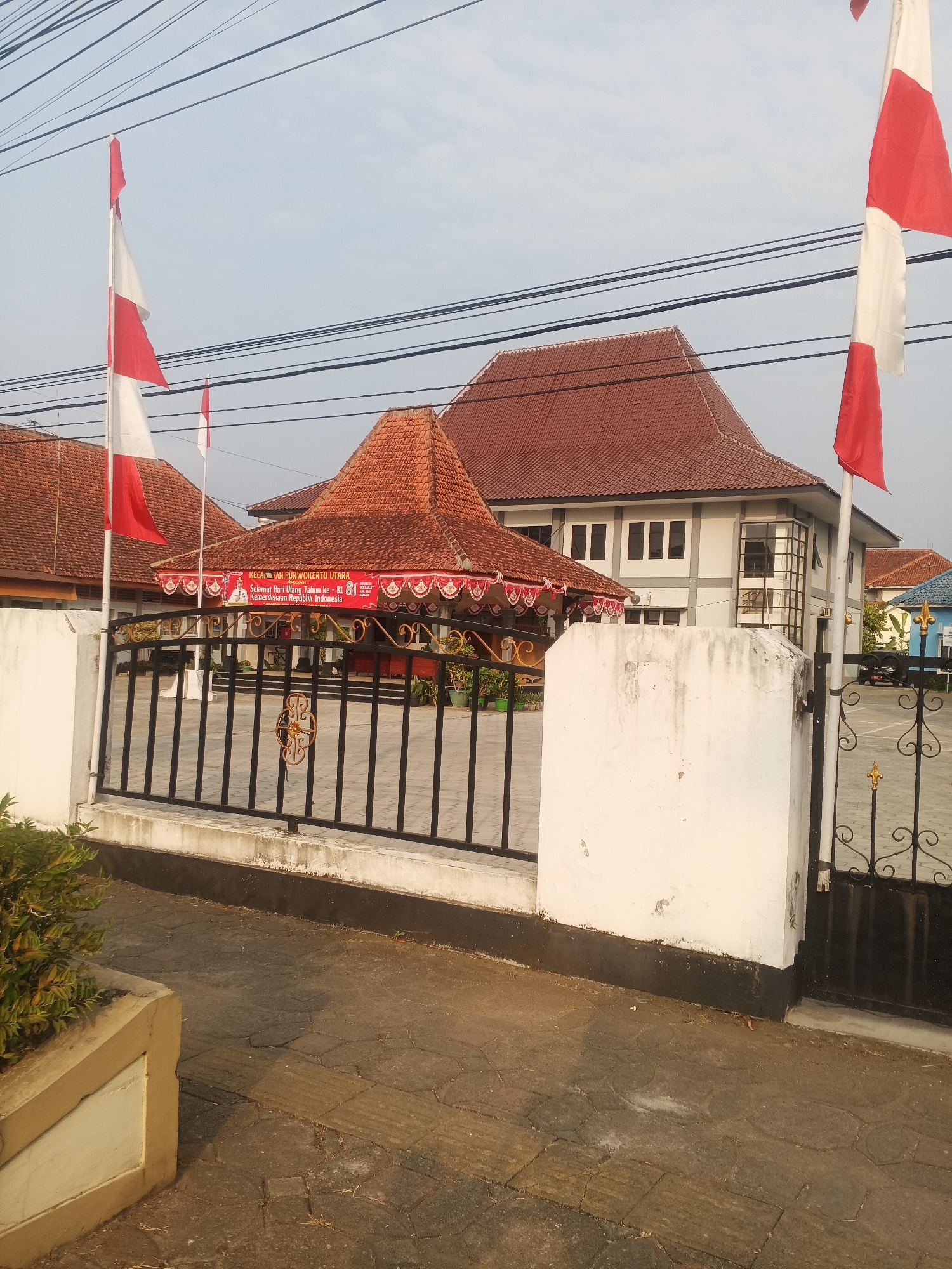 The image shows the front of a building complex, likely a government or public institution, in Bancarkembar, Indonesia. The architecture features traditional Indonesian tiled roofs in a reddish-brown hue, contrasting with modern white and gray building structures. Several Indonesian flags, red and white, are prominently displayed on flagpoles.
In the foreground, a black metal fence with decorative elements encloses the area. A white concrete pillar is visible to the left of the fence. The ground inside the fenced area appears to be paved with interlocking blocks. A banner with Indonesian text and an image of a person is stretched across the front of a covered area within the complex. The banner reads "KECAMATAN PURWOKERTO UTARA" and "Selamat Hari Ulang Tahun ke - 81 Kemerdekaan Republik Indonesia".
The environment suggests a clear sky with some visible power lines crisscrossing overhead. The lighting indicates it is likely daytime, possibly morning or late afternoon given the shadows. There are no people clearly visible in the image. The overall scene conveys a sense of civic pride and celebration, likely related to Indonesia's Independence Day.