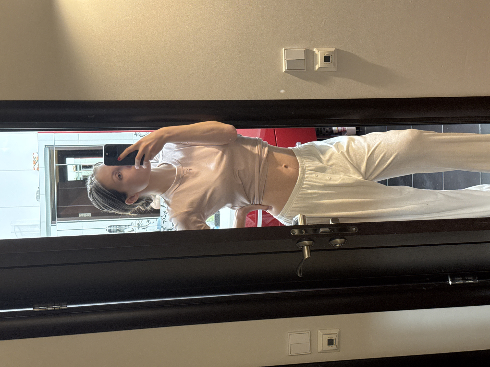 A woman is seen in a mirror selfie. She is wearing a light-colored t-shirt and white sweatpants. Her hair appears to be styled with streaks of a darker color. The background reflection shows a partial view of a room, including what appears to be a glimpse of a red object and other furniture. The reflection is contained within the frame of a dark-colored door. Wall features above and below the door frame suggest an interior setting. The location is not discernible as Moscow, Russia without further information.