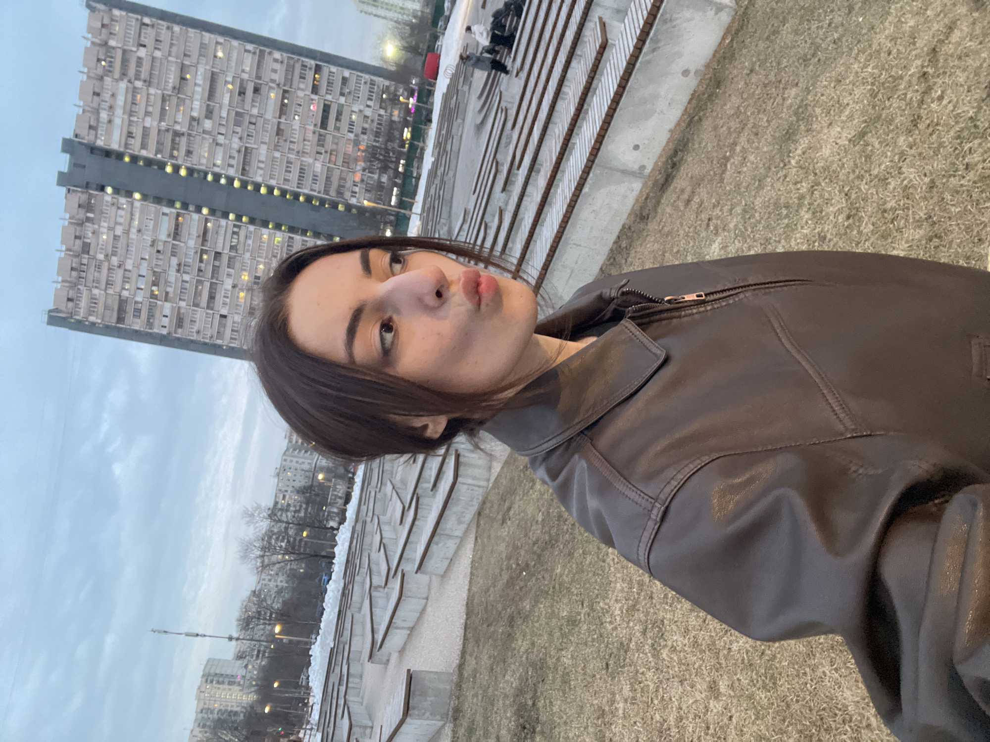 The image depicts a young adult female, partially visible in a selfie-style shot, wearing a dark brown leather-like jacket with a zippered front. She is making a puckered lip expression and looking towards her right.
The scene is an urban park, identified as Khodynka Field Park in Moscow, Russia. The ground in the immediate foreground consists of dry, sparse grass. Behind the subject and extending into the midground is a large, terraced amphitheater-style seating area constructed from concrete blocks topped with wooden planks. Two indistinct figures are visible further back within this seating area on the right side. Beyond the amphitheater, patches of snow are visible on the ground, with bare trees scattered throughout.
In the background, several multi-story residential buildings are prominent, including a tall, narrow high-rise with a distinctive dark central vertical segment and numerous uniformly arrayed windows and balconies, characteristic of the Grand Park residential complex adjacent to Khodynka Field Park. The sky overhead is overcast with light gray clouds. The overall lighting suggests late afternoon or early evening.