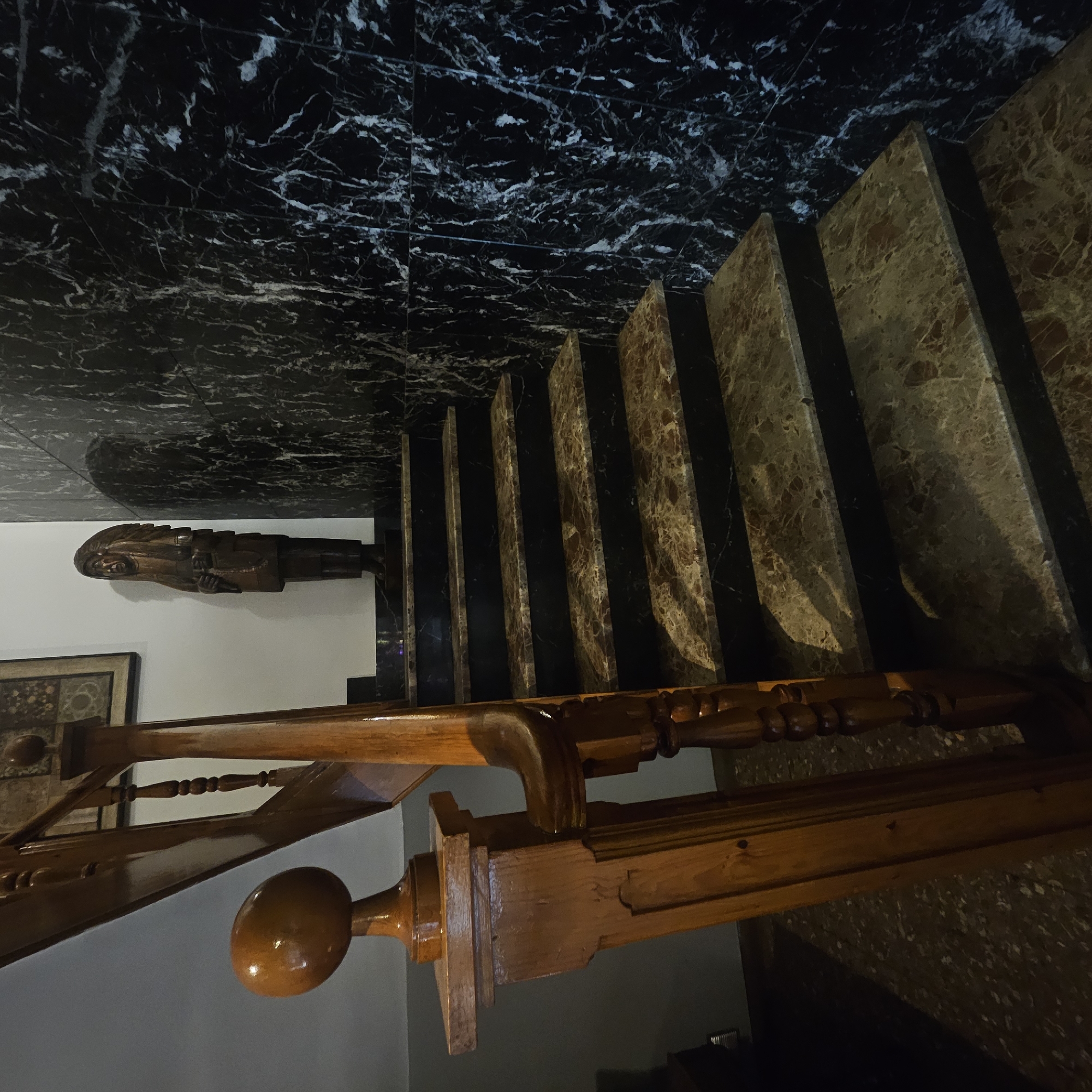 The image shows a wooden staircase with marble steps and a marble wall. A wooden statue of a Native American chief is standing on one of the steps. The staircase is lit by ambient light, casting shadows on the steps. The location of Almadrones, Spain is not visible in the image.