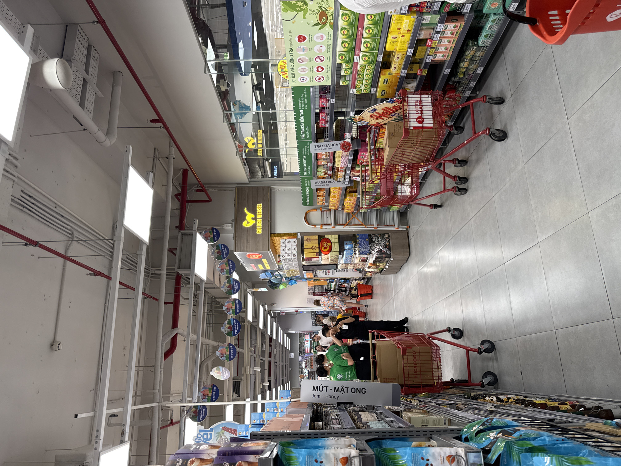 This is a wide-angle, eye-level shot inside a brightly lit grocery store in Nha Trang, Vietnam. The store appears to be a supermarket or hypermarket, featuring rows of shelves stocked with food products and beverages. The flooring is a light-colored tile.
In the foreground, to the left, are aisles of merchandise. A red shopping cart is prominently positioned in the center of the frame, filled with boxes and items. To the left of the cart, a sign with Vietnamese text is visible, likely indicating a product category. Behind the cart, several store employees wearing green shirts are interacting with a customer or fellow employee dressed in black. Further back, other shoppers can be seen browsing the aisles.
The shelves are organized with various packaged goods, including boxes of tea and other food items. Above the shelves, signage and decorative elements are visible, including circular hanging signs with text and graphics, and larger banners with product advertisements. The ceiling shows a complex network of pipes, conduits, and lighting fixtures. One prominent sign, featuring a yellow weasel graphic, reads "GOLDEN WEASEL."
The overall atmosphere suggests a typical, busy shopping environment.