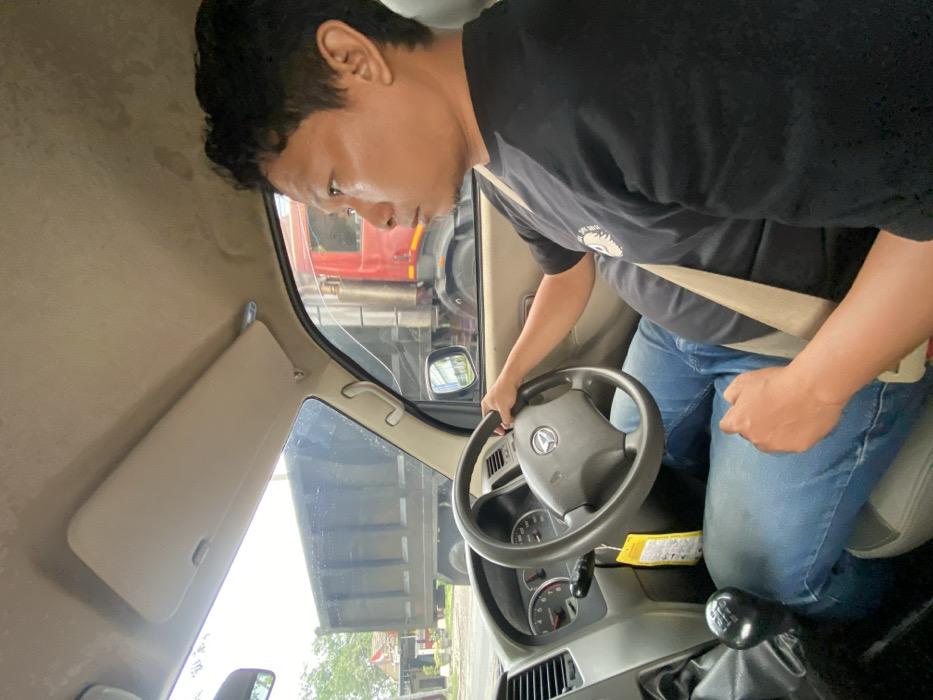 An Asian man wearing a black t-shirt and blue jeans is seated in the driver's seat of a truck. He has a beige seatbelt fastened across his chest. He is looking towards the windshield with his hands on the steering wheel. The interior of the truck is a light beige color. Through the windshield, a red truck is visible. The sun visor is down.
