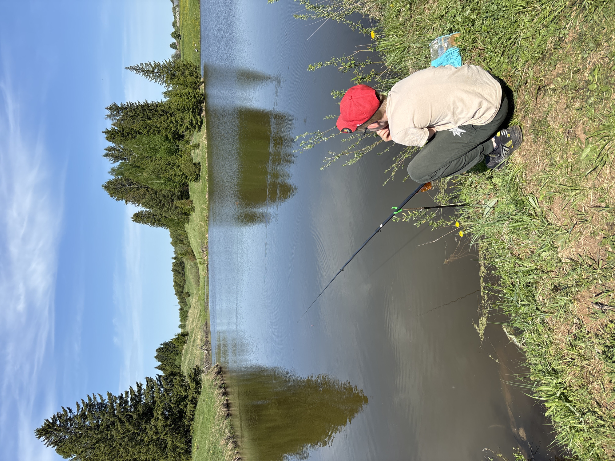 A young individual, likely a child, wearing a red baseball cap, light-colored shirt, and dark pants is crouching by the edge of a body of water, possibly a pond or lake. They are holding a fishing rod, with the line extending into the water. The setting appears to be a rural area with grassy banks along the water's edge and trees lining the opposite side. There are some small yellow flowers and vegetation near the water. A box of fishing bait is lying on the grass next to the child. The location is Karashur, Russia.