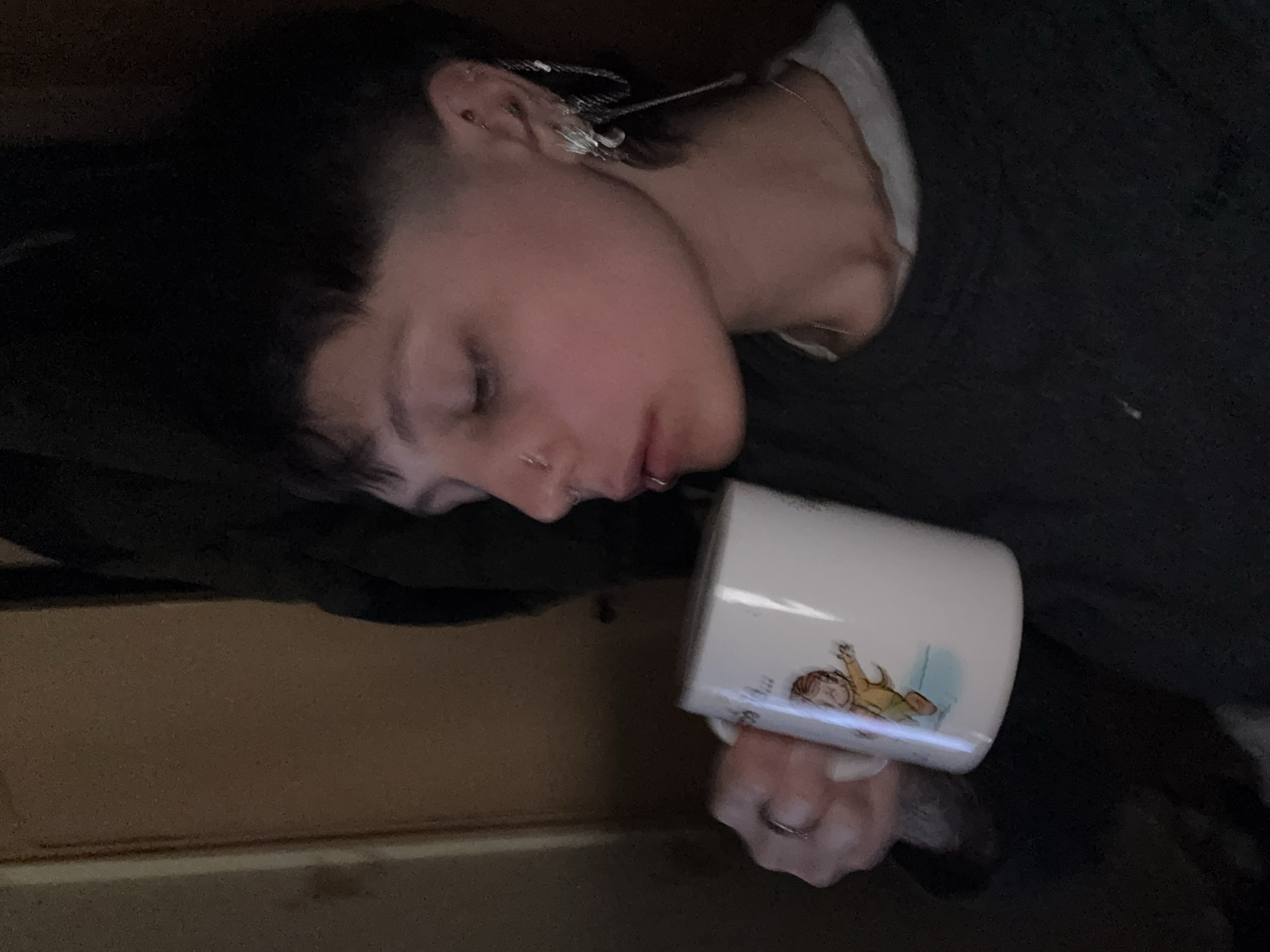 A person with a shaved head, multiple piercings in their left ear, and a nose ring is seen lying down, apparently asleep. They are wearing a dark top and a white undershirt. They are holding a white mug with a cartoon-style illustration of a person on it. The location is not discernible from the image, so the claim of being in Moscow, Russia cannot be confirmed. The background consists of dark, indistinct surfaces and a portion of what seems to be a wall or furniture. The person appears to be in a state of repose while holding the mug.
