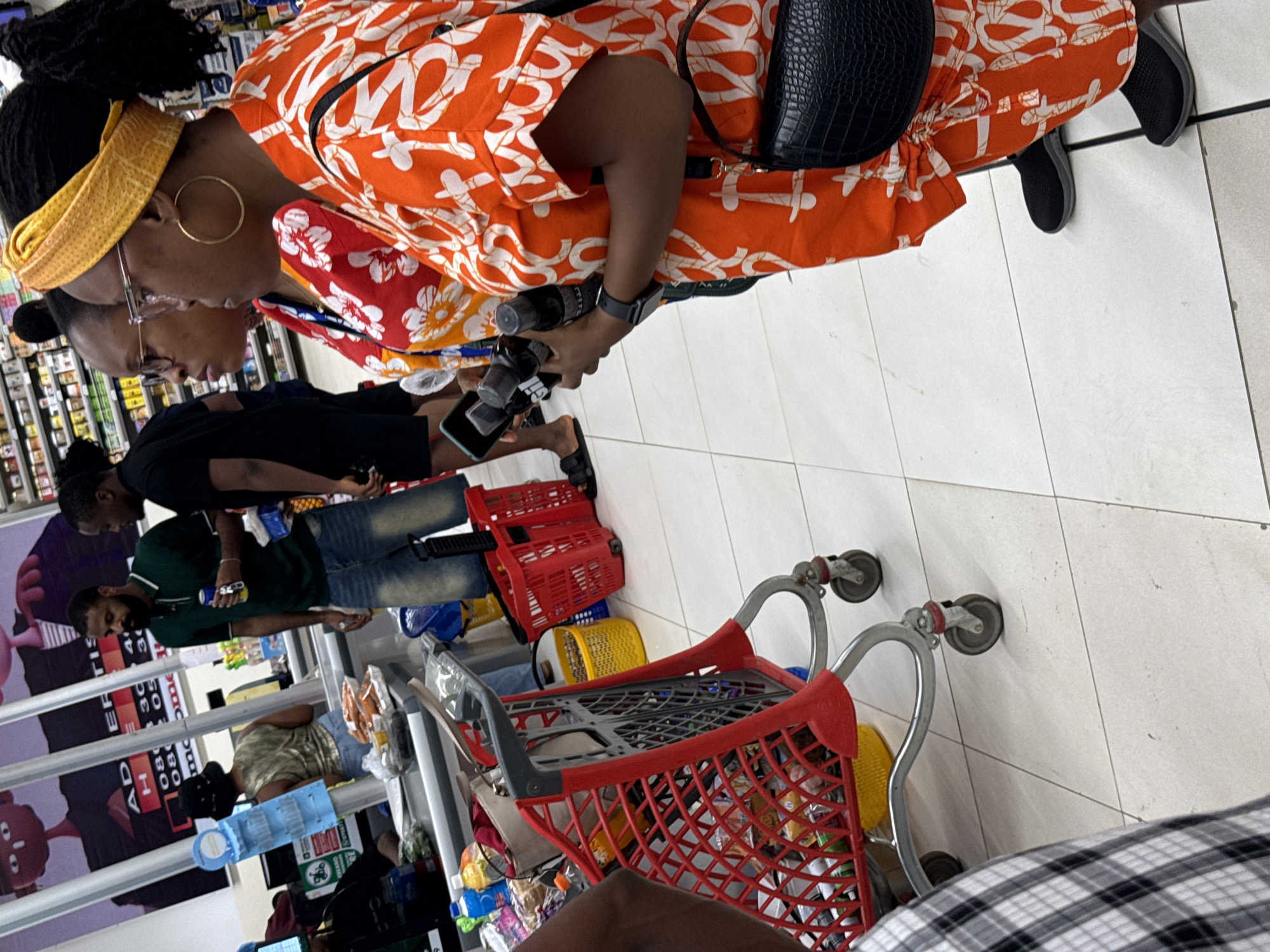 The image depicts the interior of a well-lit supermarket in Abuja, Nigeria. The floor is tiled with light-colored square tiles.
Several individuals are visible in the scene. A woman with her hair styled in a bun and adorned with a yellow headband wears glasses and large hoop earrings. She is dressed in a vibrant orange and white patterned top. A black crocodile-skin bag is slung over her shoulder. She appears to be holding small bottles and a smartphone.
To her left, another woman also wears glasses. Both women are partially obstructed by shopping carts and other shoppers.
Further down the aisle, a man in a green shirt and jeans is visible, along with other shoppers, including one in a black shirt.
A red shopping cart is prominently displayed in the foreground, filled with various items. There are also shopping baskets of various colors (red, yellow, and blue) stacked nearby. The store's shelving units are stocked with diverse products. A promotional banner featuring cartoon characters is visible on the left side of the frame.