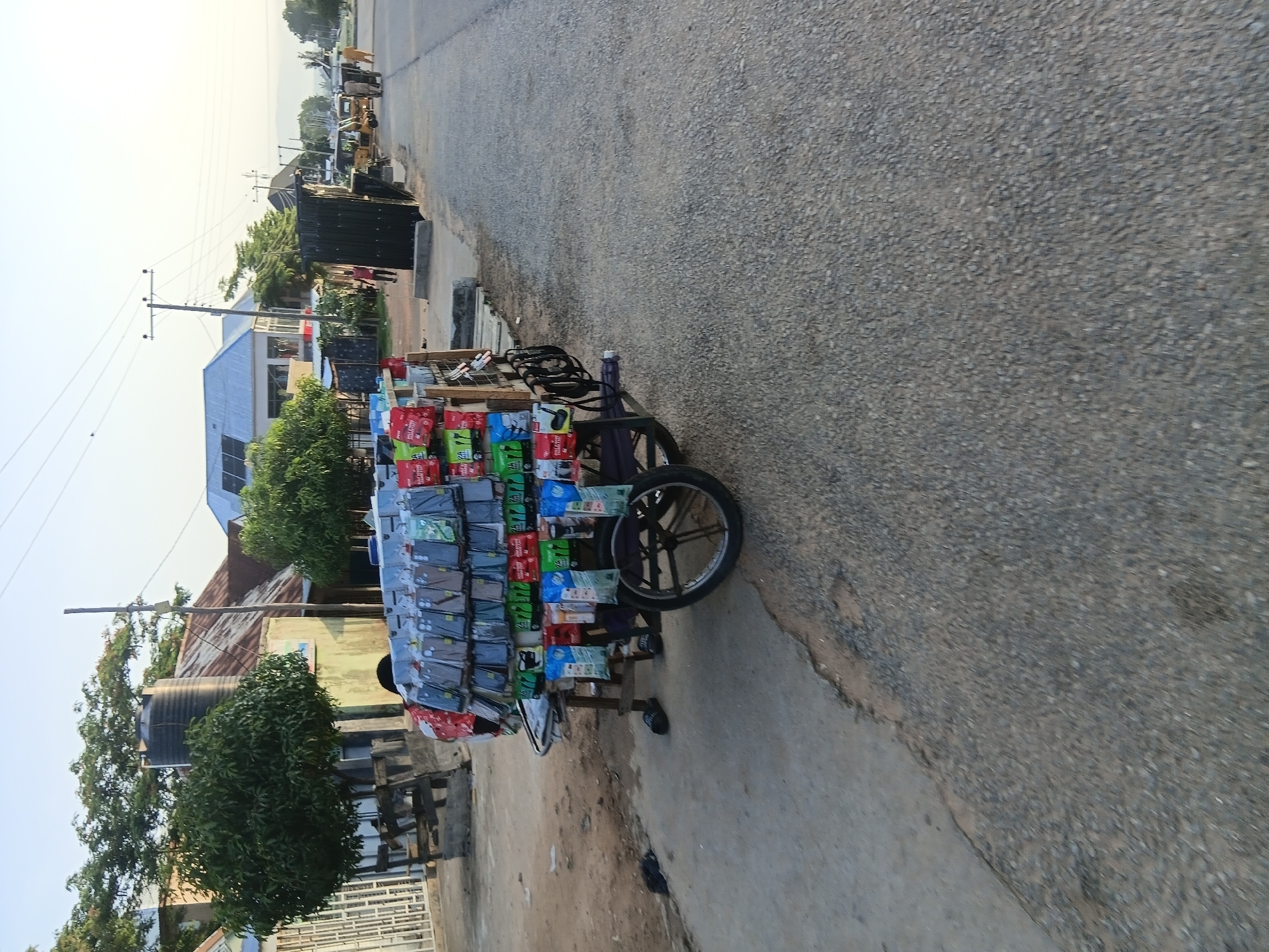 The image depicts a street scene in Jalingo, Nigeria, on a clear day with a bright sky. In the foreground, a wooden cart laden with cell phone accessories, primarily phone cases and earphones, is parked on the side of a paved road. The cases are displayed on multiple shelves, showcasing a variety of designs and colors. Some packages of earphones are also visible. The cart has two large spoked wheels, suggesting it's designed for mobile vending. Two pairs of black sandals are placed on the ground near the cart.
To the left of the cart, a dirt path leads towards a building with a corrugated metal roof and a large black water tank on top. Trees with lush green foliage surround the building and the area. A white metal fence runs along the property line.
Further down the road, a few other structures are visible, including one with a modern-looking silver roof and solar panels. People are seen walking in the distance, and a yellow construction vehicle and a motorbike are also present on the road. The overall environment suggests a moderately developed urban or semi-urban area. The scene appears to be a typical street-level marketplace in Jalingo, Nigeria.