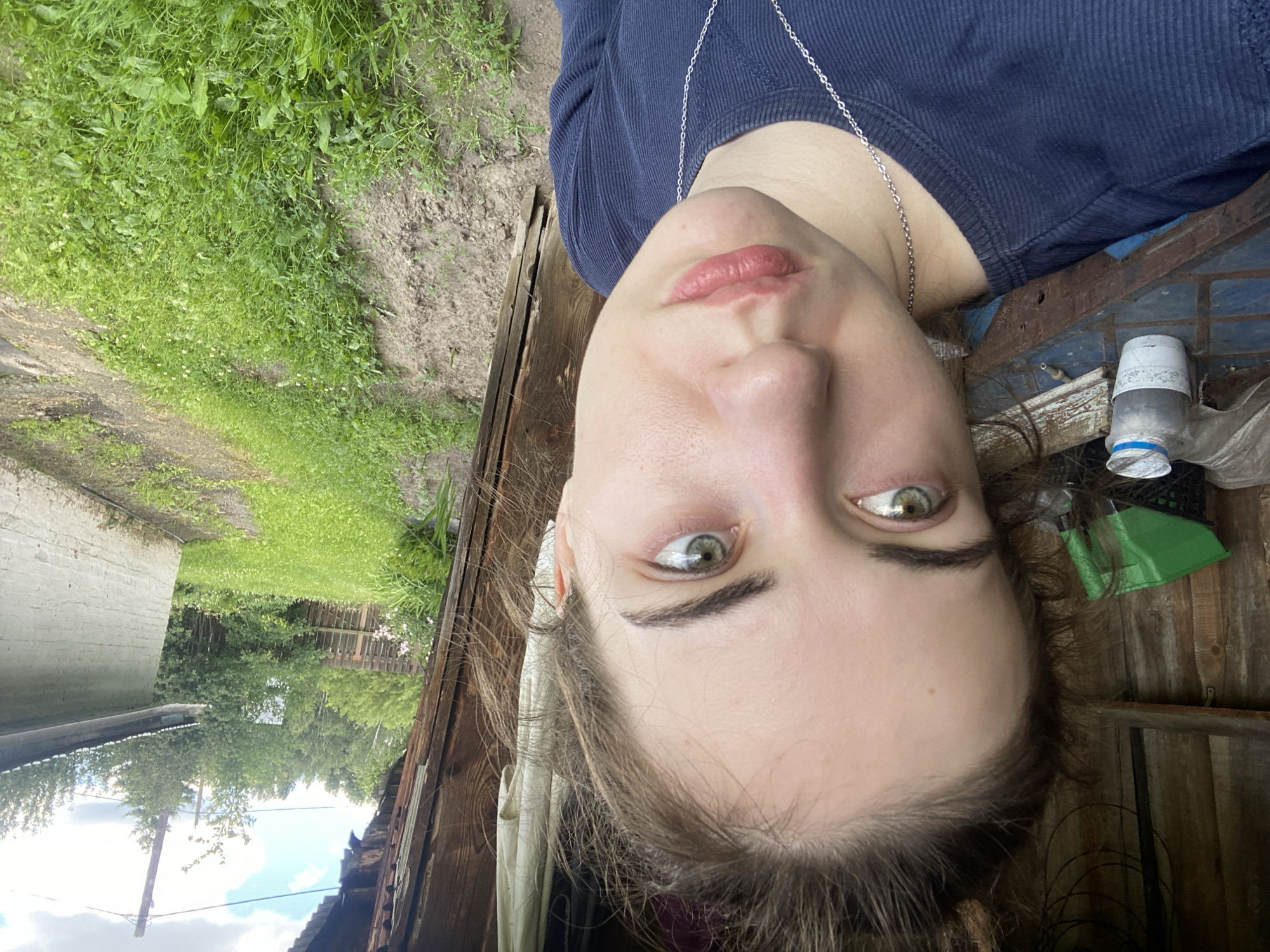 A medium close-up selfie captures a young woman with fair skin and light brown hair pulled back from her face. She has green eyes and is looking slightly to her right with a neutral expression. Her lips are slightly parted. She is wearing a dark blue long-sleeved shirt with a silver chain necklace.
The background suggests a rural or semi-rural setting, possibly in Zavodoukovsk, Russia, given the context. To the left, a wooden structure with vertical planks is visible, with a shelf holding a green plastic bin and a white container with a blue lid. Further to the left, part of what appears to be a wooden table with a blue surface is present.
To the right of the woman, a dirt path or yard is covered with green grass and weeds. In the distance, a wooden fence separates the yard from trees and foliage. A light-colored, brick-like structure with a metal roof is also visible to the right, along with a utility pole and wires against a cloudy sky. The overall lighting suggests it is daytime, with overcast conditions.
