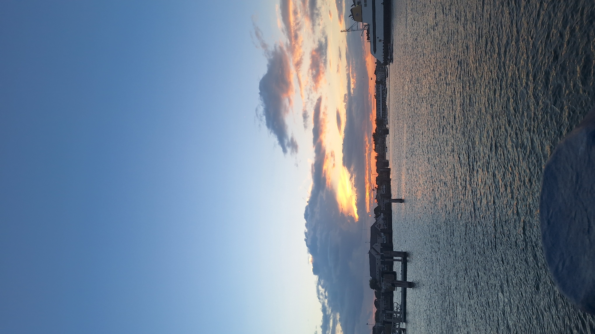A waterfront scene at sunset in Nantucket, USA, is captured in this vertical, full-frame image. The sky is a gradient of clear blue at the top, transitioning to pale orange and pink hues near the horizon, where scattered clouds are illuminated by the setting sun. The water in the foreground is a dark, rippling blue-gray, reflecting the colors of the sky. Along the distant shoreline, there is a collection of buildings with pitched roofs, characteristic of a coastal town. A ferry, painted white with a yellow stripe and the words "THE NANTUCKET" visible on its side, is docked on the right side of the frame, with a small boat nearby. In the mid-ground, several wooden posts and docks extend into the water, suggesting a marina or harbor. The overall atmosphere is serene, with the setting sun casting a warm glow over the tranquil waters and the silhouette of the town.