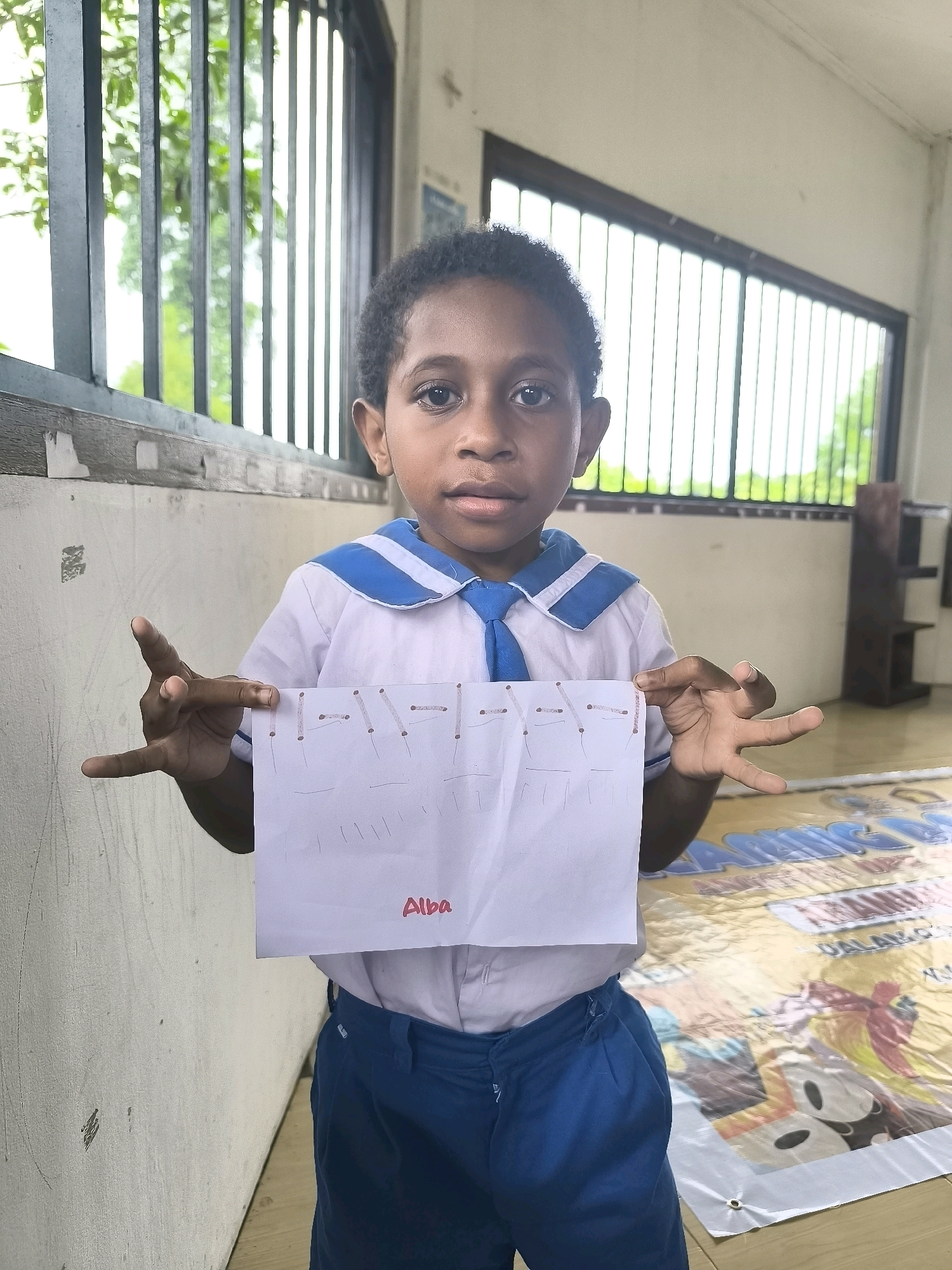 A young boy stands in front of a white wall, holding a piece of paper with drawings of letters and vertical lines. He is wearing a white school uniform with blue trim and blue pants. His left hand is positioned in a gesture that resembles the letter 'L', while his right hand holds the paper. The paper has the word "Alba" written in red on it. Behind him, a large banner with colorful cartoon characters and text in what appears to be Indonesian is visible on the floor. The environment suggests an indoor or semi-outdoor setting, possibly a school or community center.