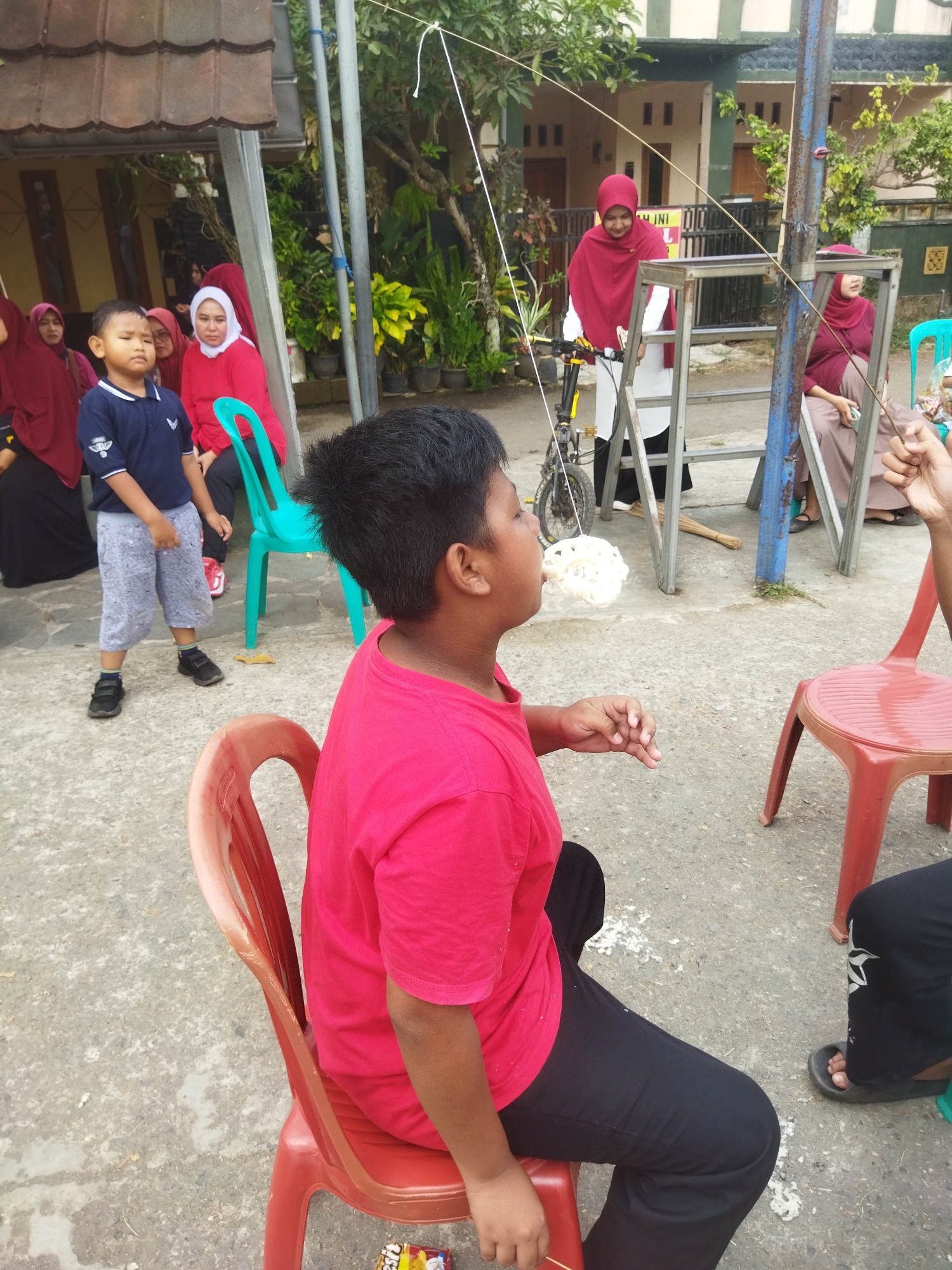In Banjar, Indonesia, a group of people are gathered outdoors for what appears to be a cracker-eating competition. In the foreground, a boy in a red shirt sits on a red plastic chair, facing right, with his mouth open, attempting to eat a cracker suspended by a string. Behind him, another boy in a blue shirt and grey shorts stands near a teal chair. Several women wearing hijabs are visible in the background, observing the event. One woman in a red hijab is standing by a bicycle and a metal frame structure. The ground is paved with concrete, and there are some scattered white specks, possibly food debris. The setting suggests a local community event or celebration.