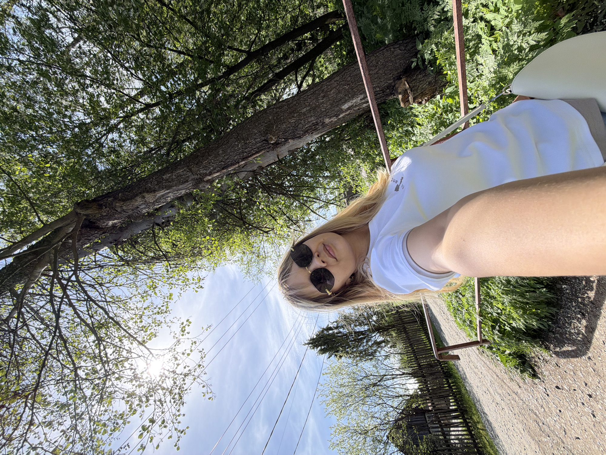 A woman with sunglasses is in Chaikovsky, Russia. She is outside, under trees and a partly cloudy sky, near power lines and a wooden fence. She wears a white t-shirt and carries a light-colored handbag. A gravel path is visible.