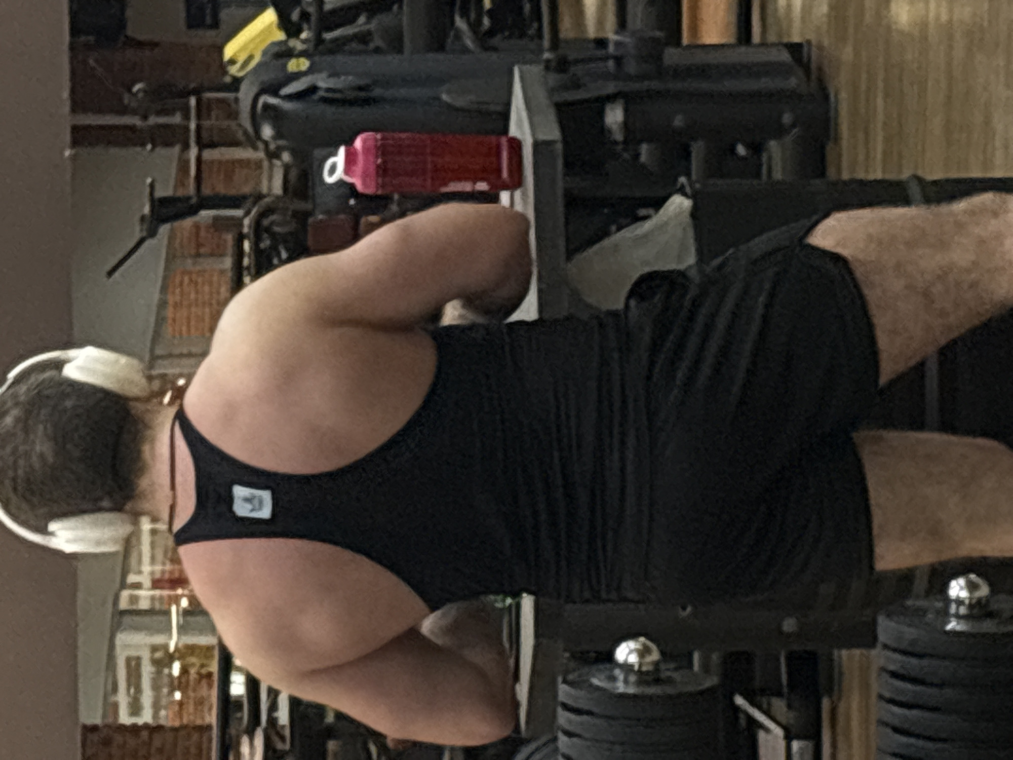 A muscular man wearing black workout attire and white headphones is seen from the back while exercising on weightlifting equipment. The setting appears to be an indoor gym. Stacks of weight plates are visible on the equipment. A red water bottle rests on a section of the equipment. The location is Chaikovsky, Russia.