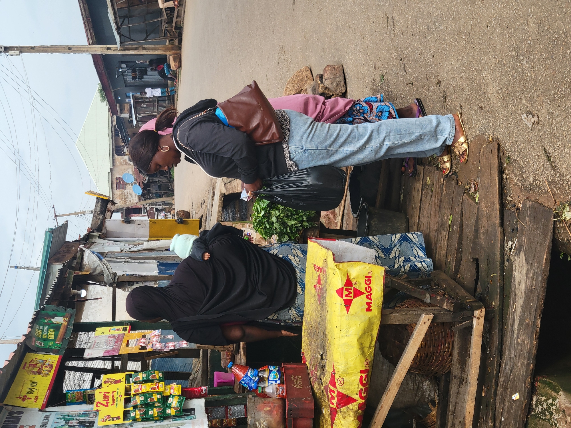 This image depicts an outdoor market scene in Jos, Nigeria. Two women are the primary subjects. The woman in the foreground, on the left, is wearing a black hijab and appears to be tending to a stall. She is partially obscured by her clothing and the stall's merchandise. The second woman, on the right, is wearing a black jacket, blue jeans, and gold sandals. She is looking down and holding a black plastic bag filled with green vegetables, and has a brown shoulder bag. A large yellow MAGGI bag is prominently displayed on the stall.
The stall itself is made of rough wooden planks and is laden with various goods, including packaged food items (evident from the "MAGGI" and "Zuri Seasoning" text on packaging), bottles of beverages, and fresh produce. The setting appears to be a street or roadside market, with buildings and a paved road visible in the background. Power lines crisscross the sky. The lighting suggests it might be daytime, possibly overcast given the diffused light. The overall impression is one of a bustling, everyday market environment. The presence of the MAGGI branding on the bag and packaging is a notable detail, indicating common household products available in the market.