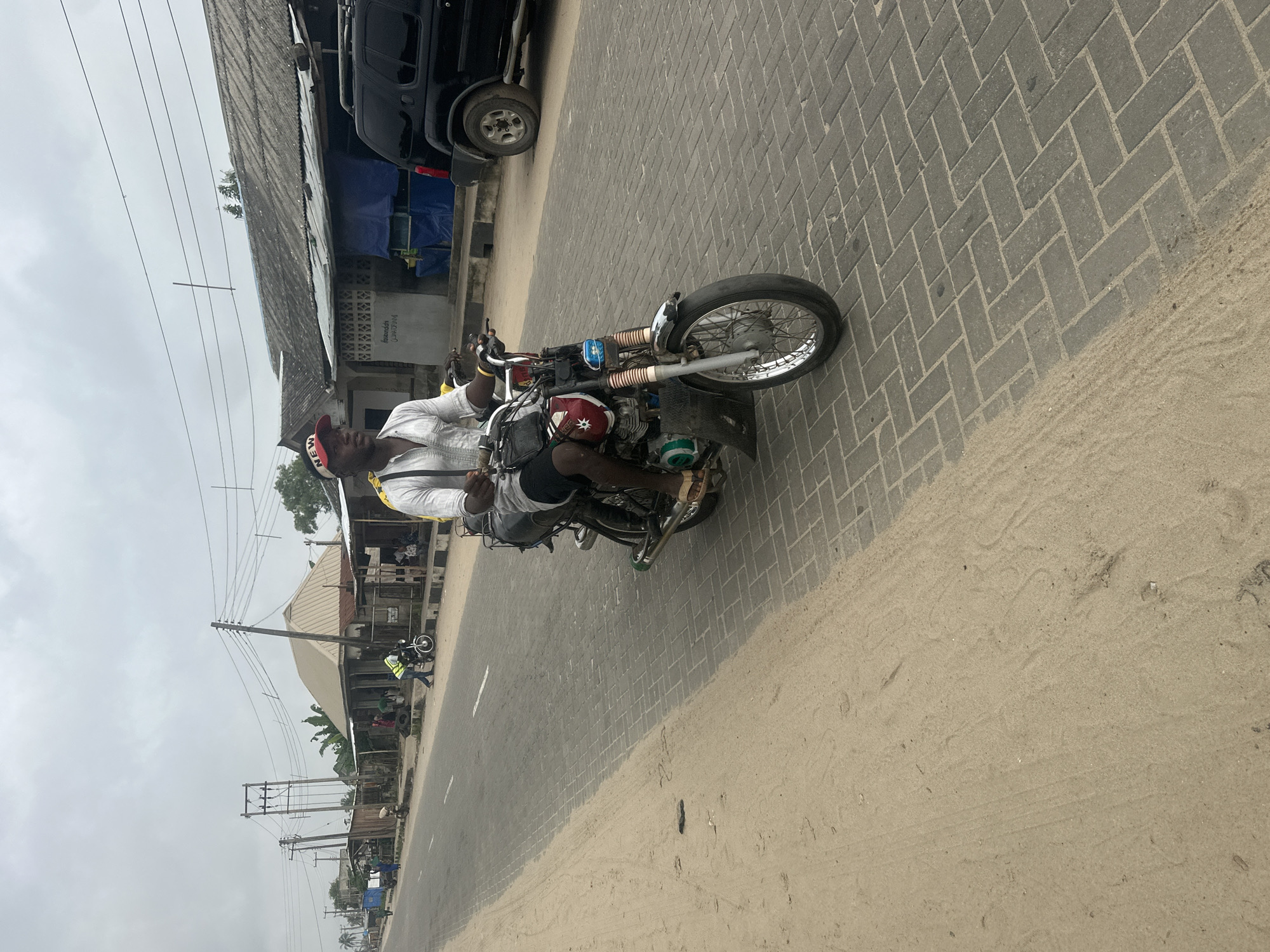 A man rides a motorcycle down a paved road in Badagry, Nigeria. The man, wearing a white shirt and black shorts, is looking forward as he drives. He has a red and white baseball cap on his head. The road is lined with buildings on the right side of the image, and there is sand on the left. The sky is overcast. A black SUV is parked on the right side of the road. The motorcycle has a silver fuel tank with red and white decals and a black seat. The motorcycle's front wheel is large and spoked. The road surface is made of gray interlocking bricks. A small, dark bag is strapped to the motorcycle's handlebars. In the background, there are power lines and poles.