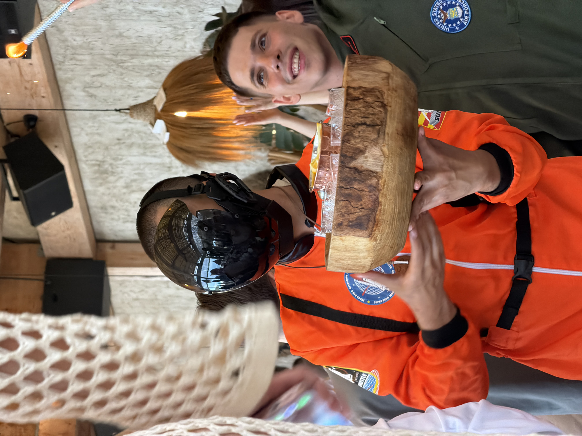 In a dimly lit interior space, two young men are captured in a close-up shot. On the left, a person wearing an orange astronaut-like jumpsuit with a black helmet that obscures their face is holding a wooden bowl filled with glassware and what appears to be fruit. The helmet has a reflective visor that shows a distorted reflection of the surroundings, including a ceiling and other people. The jumpsuit has patches on the left arm, one of which is partially visible and circular with text and imagery. On the right, another young man in a green military-style uniform, possibly an Air Force uniform given the patch on his chest, looks upwards and to the right with a wide smile, his mouth open as if speaking or laughing. His uniform features a circular patch with the United States Air Force insignia. The background reveals details of the venue, including a textured wall, wooden beams, and a decorative straw-like lampshade hanging from the ceiling, lit from within by an orange light. The overall atmosphere appears to be casual and celebratory.