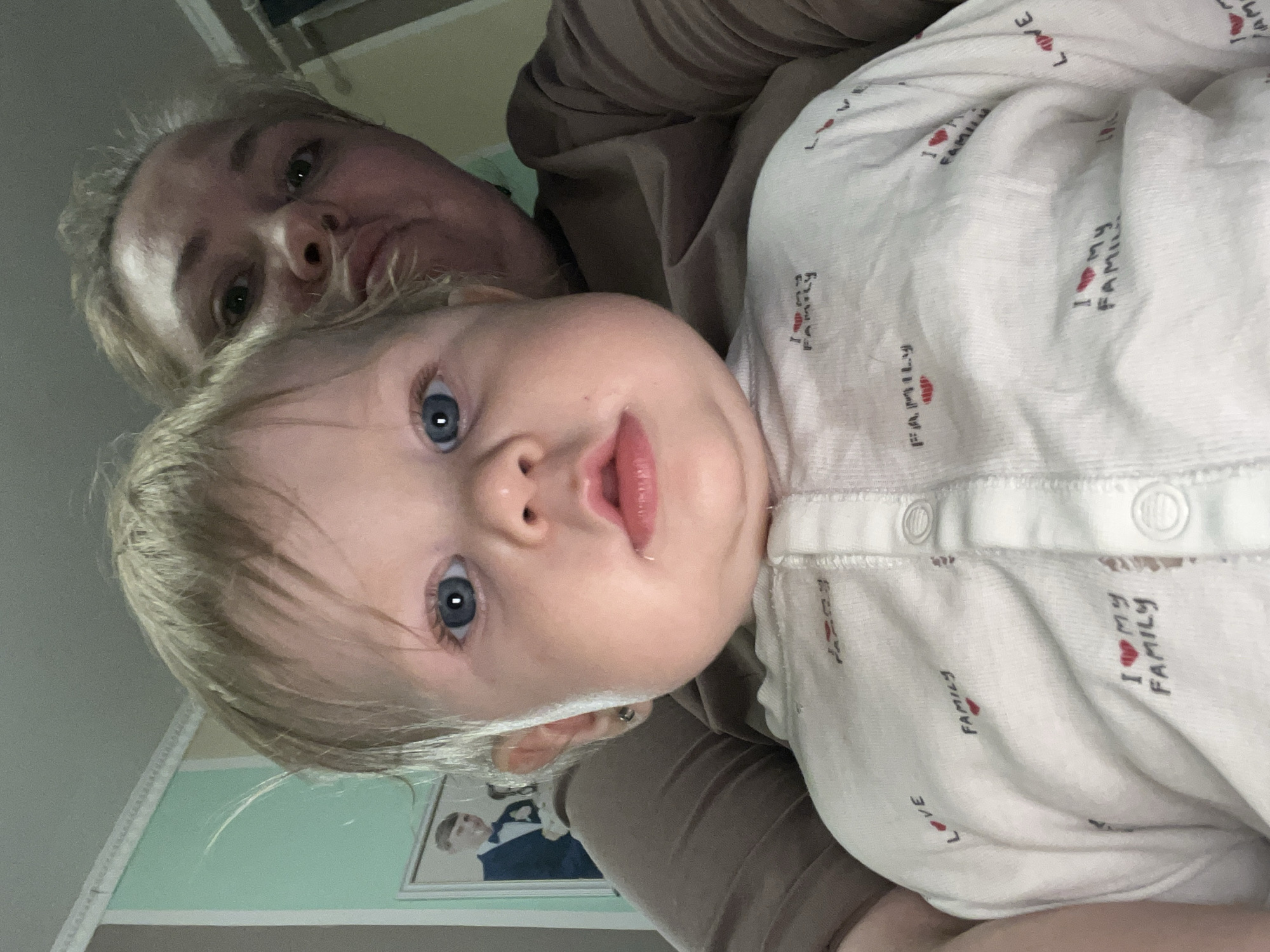 A selfie shows a fair-skinned adult woman and a fair-skinned baby, likely related. The baby is wearing a white onesie printed with the phrase "I love my family" and has light blond hair and blue eyes. The woman has blond hair and is wearing a brown sweater. A framed photograph is visible on the light green wall in the background. The location is likely a residence in the city of Oryol-Izumrud, Russia based on the user's query.