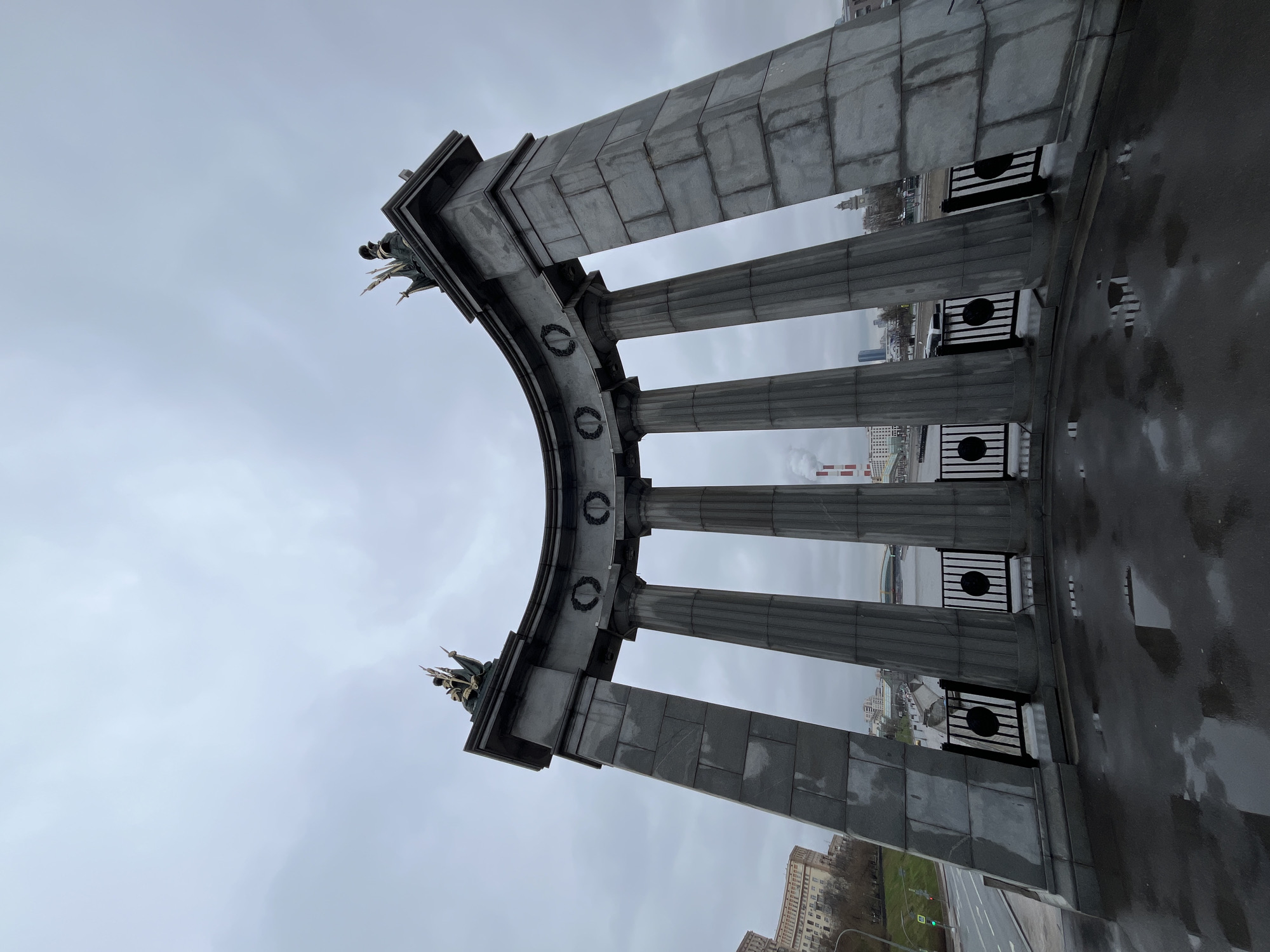 The image depicts the Poklonnaya Hill Victory Park in Moscow, Russia. The scene shows a curved monument with classical columns and wreaths. The sky is overcast. The wet ground reflects light, indicating rain. In the background, buildings and industrial structures are visible. No people are identifiable.