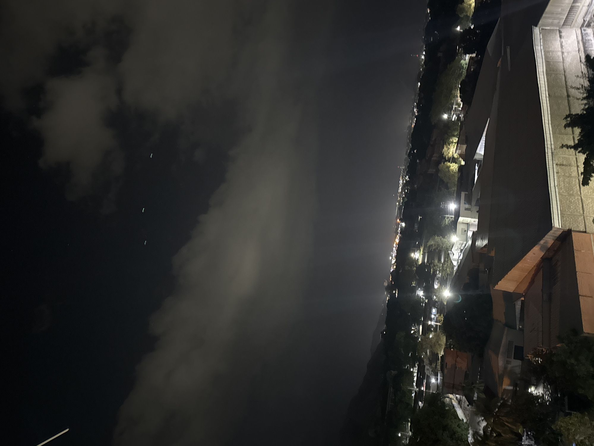 At night, the city of Maracay, Venezuela, is visible from an elevated viewpoint. The scene is dominated by the city skyline stretching along the right side of the frame, with buildings, streetlights, and other light sources creating a line of illumination. To the left, the sky is overcast with heavy clouds. The angle of the photograph suggests it was taken from a high location, perhaps a building or hillside. There are no people visible.