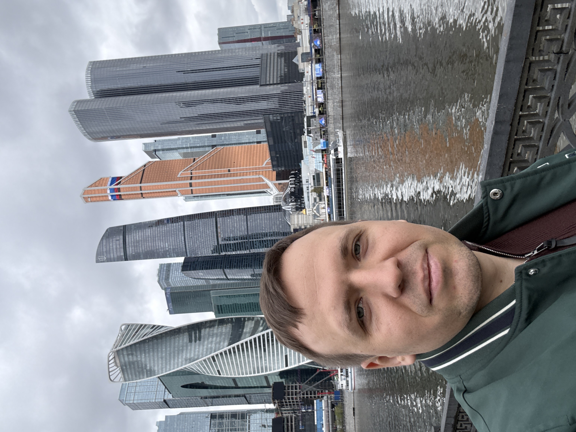 A man takes a selfie with the Moscow International Business Center (Moscow-City) skyline in the background. The skyline features several modern skyscrapers of varying shapes and heights, reflected in the river. The weather appears overcast. The man is wearing a green jacket with a striped collar and a maroon undershirt. He stands along a stone or concrete embankment with decorative metal railings. The water reflects the sky and the buildings.