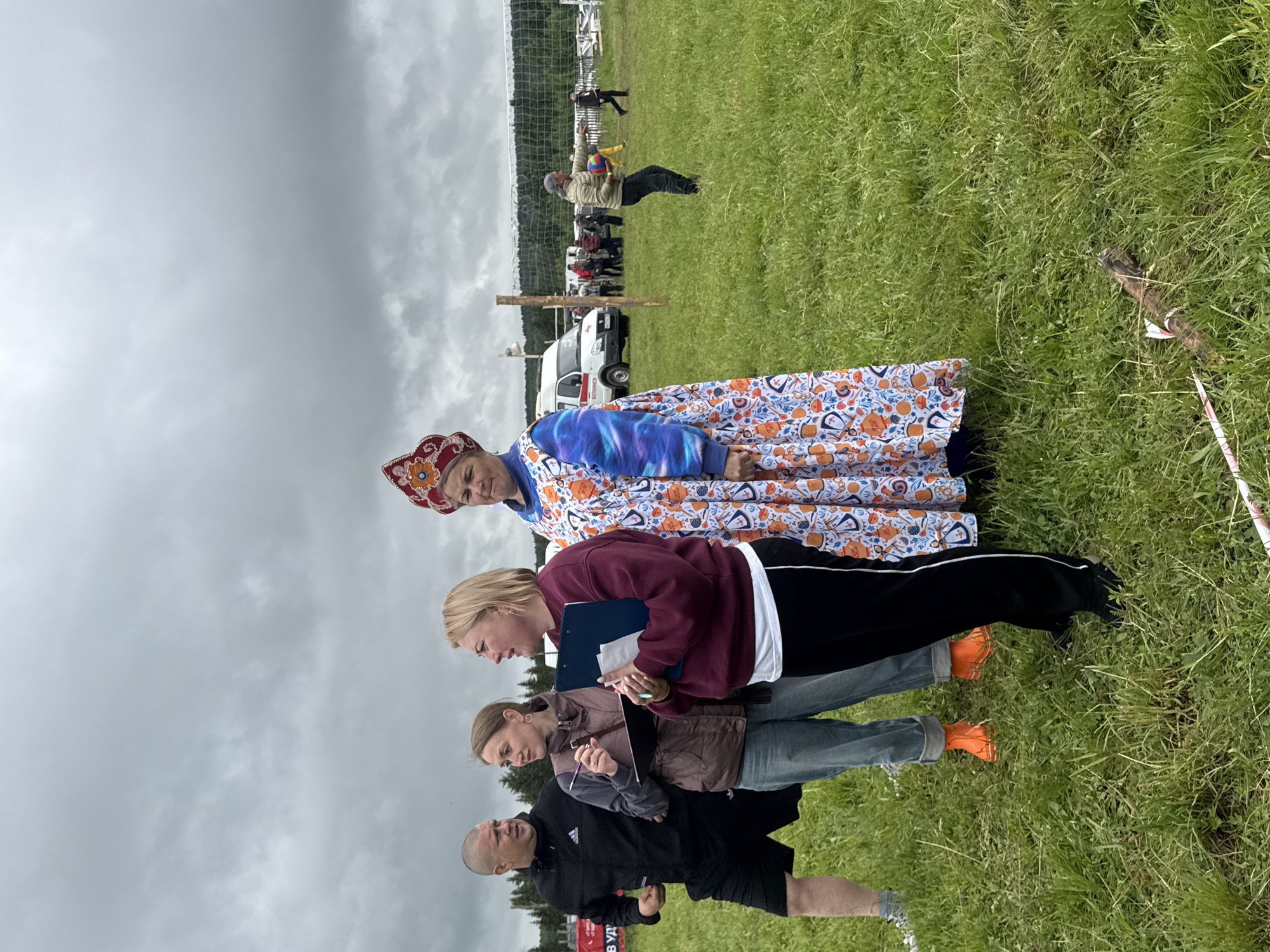 This image depicts an outdoor scene in Большая Кивара, Russia, under an overcast sky, likely at a community event or festival. The foreground is dominated by a group of four individuals standing on a patchy green grassy field.
From left to right:
1. A bald or closely shaven man in a black Adidas tracksuit top and black shorts stands with his left leg forward, appearing to be in motion or about to move. He looks towards the two women next to him.
2. A woman with brown hair, wearing a brown quilted vest over a long-sleeved top and light blue jeans, holds a dark clipboard and a pen, looking towards the right. She has orange rubber boots on her feet.
3. A blonde woman in a burgundy sweatshirt, black pants, and a white undershirt holds a blue clipboard and a pen, looking and speaking towards the right, her mouth slightly open. She wears black shoes.
4. A woman in traditional Russian attire stands partially visible on the far right. She wears an elaborate red kokoshnik-style headdress with gold and floral embellishments, and a long, light-colored dress or sarafan with a vibrant pattern of orange, blue, and white elements. Her left arm has a blue and purple tie-dye pattern sleeve. She looks straight ahead with a neutral expression.
In the midground, behind these individuals, a white ambulance with red crosses and Cyrillic text is parked. To the right of the ambulance, a volleyball net is set up between two wooden poles, with several people visible beyond it, some appearing to be playing volleyball. One man in a light jacket is visible holding a colorful volleyball and pointing to the right. A dense line of evergreen trees forms the horizon in the background, beneath the uniform gray, cloudy sky. On the ground in the foreground, near the right edge, a short wooden log or branch lies horizontally on the grass, with a piece of pink and white tape or ribbon attached.