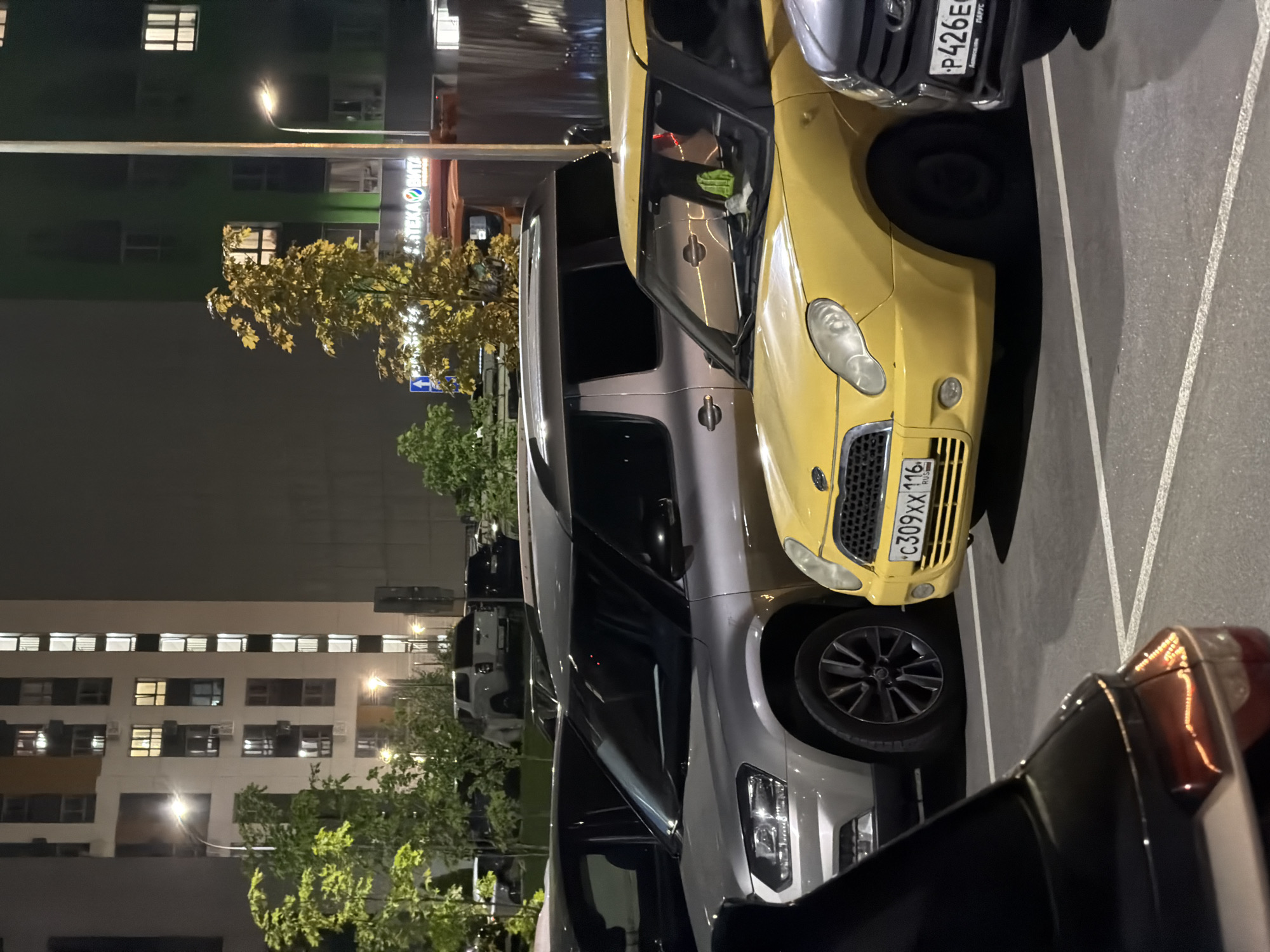 In an outdoor parking lot at night, several vehicles are parked in front of a modern apartment building. The building is illuminated with numerous windows lit from within, and a green-colored section is visible on the right side. Trees with green and yellow foliage are scattered throughout the scene. A streetlamp casts light on the parking area.
The foreground features the rear of a black car, partially obscuring a silver SUV in the middle ground. To the right of the silver SUV, a yellow compact car is parked, displaying a license plate with Cyrillic characters and the text "C309XX 116 RUS". Further to the right, a dark-colored vehicle is visible with a license plate showing "P426EC". In the background, behind the silver SUV, other vehicles are parked, including a white pickup truck. A tall fence separates the parking area from some structures in the far background. The scene suggests a residential urban environment.