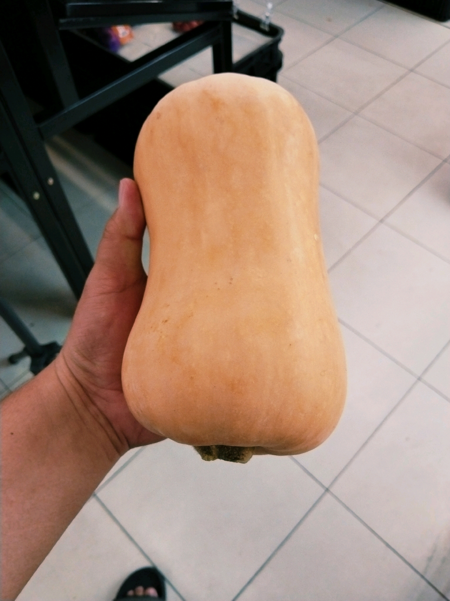 In Surabaya, Indonesia, a person's hand holds a butternut squash. The squash is light orange and has a smooth texture. It is positioned centrally in the frame. In the background, a black metal structure is visible, suggesting it might be shelving or furniture. The floor is tiled with light-colored square tiles, indicative of an indoor setting, possibly a market or grocery store. A person's feet wearing black sandals are partially visible at the bottom of the frame.