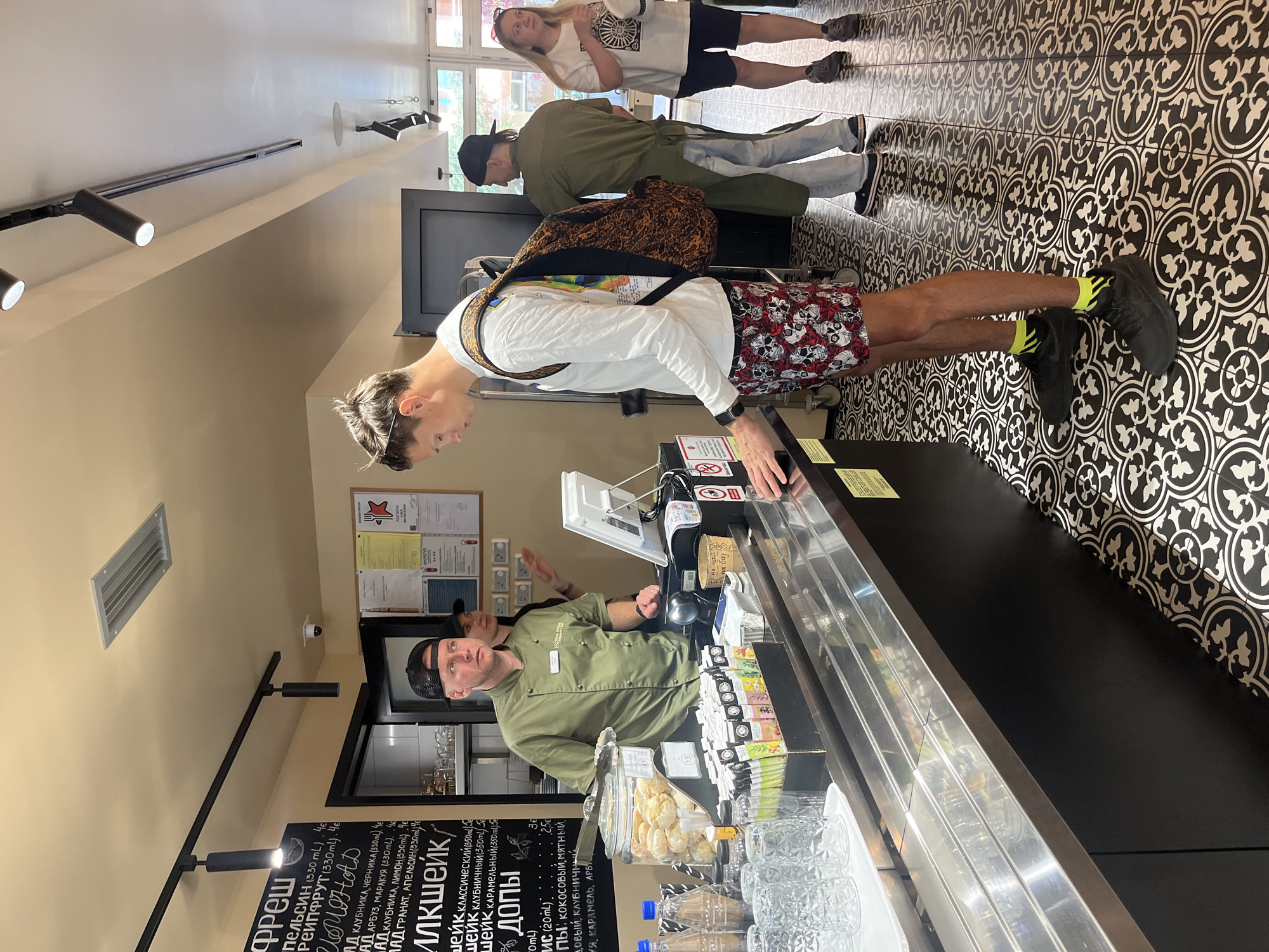 The image captures the interior of a brightly lit cafe or quick-service restaurant. In the foreground, a young man with light brown hair, wearing a white long-sleeved shirt, skull-patterned shorts, black sneakers with neon yellow socks, and a colorful backpack, stands at a black counter. He is leaning forward, looking at the items displayed or interacting with the staff.
Behind the counter, a male staff member in a green chef's jacket and a black cap is visible, looking towards the customer. On the counter, there's a cash register, a screen, a card reader, a glass jar filled with cookies or pastries, clear drinking glasses, and several bottled drinks. Various packaged items, possibly tea bags, are also arranged on the counter.
Further back in the line, another person wearing a green top, light-colored jeans, and a black cap is partially visible. Behind them, a woman with long blonde hair, dressed in a white t-shirt and black shorts, stands with her arms crossed.
The floor features a distinctive black and white patterned tile design. The walls are a light neutral color, and track lighting runs along the ceiling. A large window in the background suggests it is daytime. A menu board on the wall lists various fresh drinks and milkshakes in Cyrillic, with prices and sizes. Visible text includes "Фреш" (Fresh), "Милкшейк" (Milkshake), and flavors like "клубника" (strawberry), "лимон" (lemon), "карамель" (caramel), and "апельсин" (orange), along with "ДОПЫ" (add-ons) like "кокосовый" (coconut) and "мятный" (mint). Patrons appear engaged in the ordinary activity of ordering or waiting in line at the establishment.