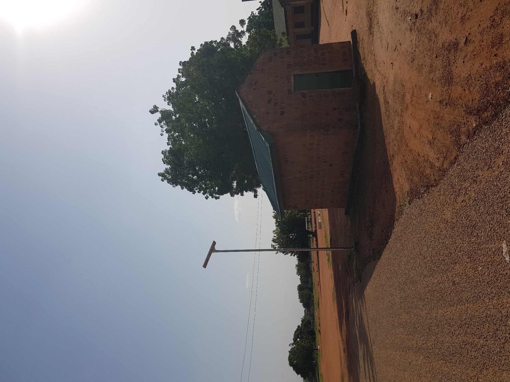 The image depicts an outdoor scene in Jimeta, Nigeria. The primary subjects are a brick building with a green door, a large leafy tree, a tall pole with a solar panel, and a dirt path. In the background, more trees and another building are visible. The setting appears to be a school or a communal area, indicated by the presence of the buildings and the open space.
The image is captured during the daytime, with bright sunlight casting shadows, suggesting it's either morning or afternoon. The sky is clear and pale blue, indicating fair weather. The ground is covered with reddish-brown dirt and gravel. A notable detail is the solar panel on the pole, which suggests a modern infrastructure element within the location.
No people are visible in the image, nor are any specific activities taking place. The composition is static, focusing on the architectural elements and the natural surroundings. The lighting and shadows are a prominent feature, emphasizing the open and sunny environment. The image's orientation is skewed, with the sky appearing on the left side of the frame and the ground on the right, suggesting the photo was taken from a low angle and rotated. There is no visible text in the image.