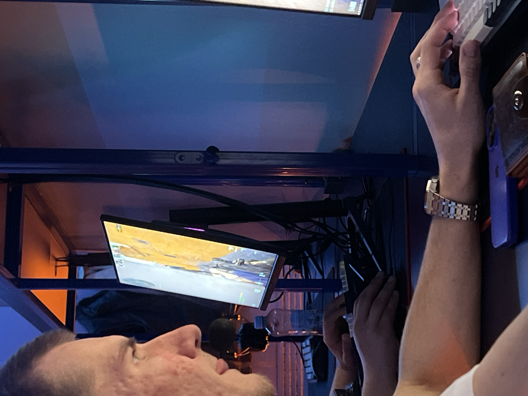 A person is lying on their back looking up at a computer screen displaying a video game. The person's head is oriented toward the lower left of the frame. A hand is on a keyboard on the upper right side. The environment appears to be indoors, with various cables and tech equipment visible above the person. The city and location are unknown.