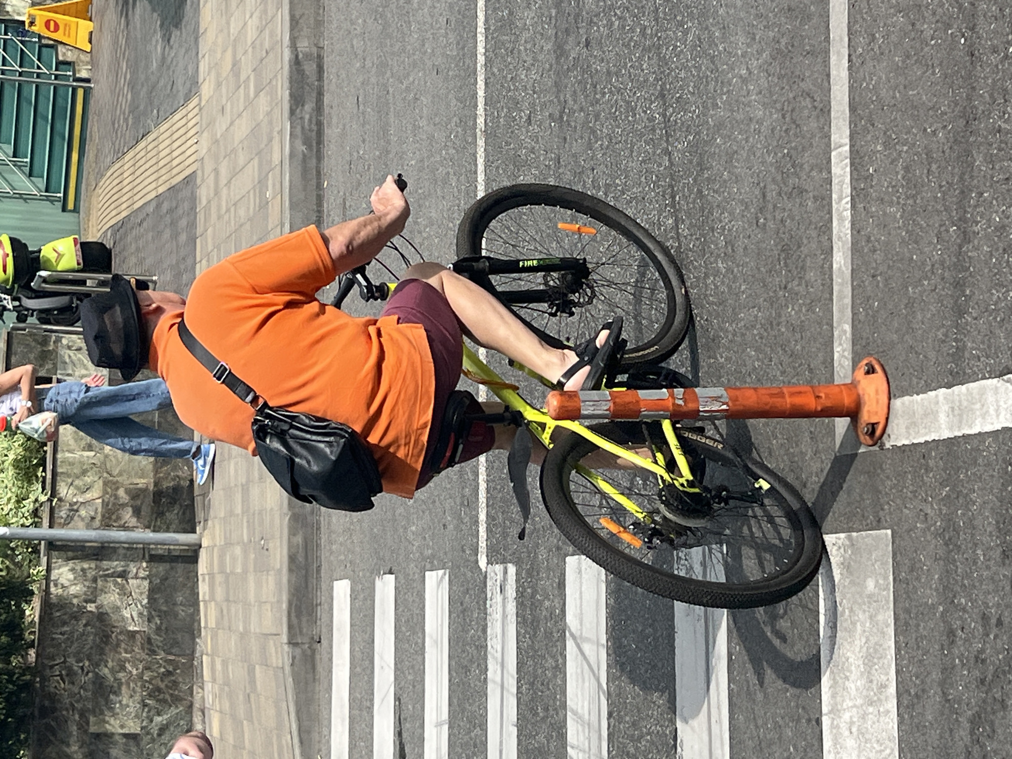A man wearing an orange polo shirt, dark shorts, and a black hat rides a yellow bicycle across a crosswalk in Sochi, Russia. He has a black messenger bag slung over his right shoulder. The crosswalk is marked with white stripes on asphalt. In the background, a woman in jeans and a patterned top walks on a sidewalk with a white railing and green stairs. A yellow scooter is parked near a yellow warning sign with a red circle. The scene is illuminated by sunlight, casting shadows.