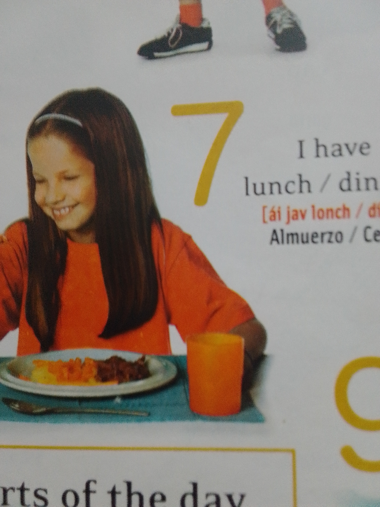 This media file features a young girl with long, dark hair and a headband, wearing an orange t-shirt, smiling as she looks down at a plate of food. The food appears to be a meal of mashed potatoes and some sort of meat. She is seated at a table with a blue placemat and an orange cup. To the right of the girl, a large yellow number "7" is prominently displayed, along with text that reads "I have lunch / din", "[ái jav lonch / di", and "Almuerzo / Ce". In the upper portion of the image, out of focus, are two pairs of feet wearing athletic shoes, suggesting a potential connection to a sports or activity context. The bottom of the image shows partial text of "rts of the day". There are no clear indicators of the time of day, weather, or specific location beyond the general context of a mealtime. The girl's expression suggests happiness and engagement with her meal.