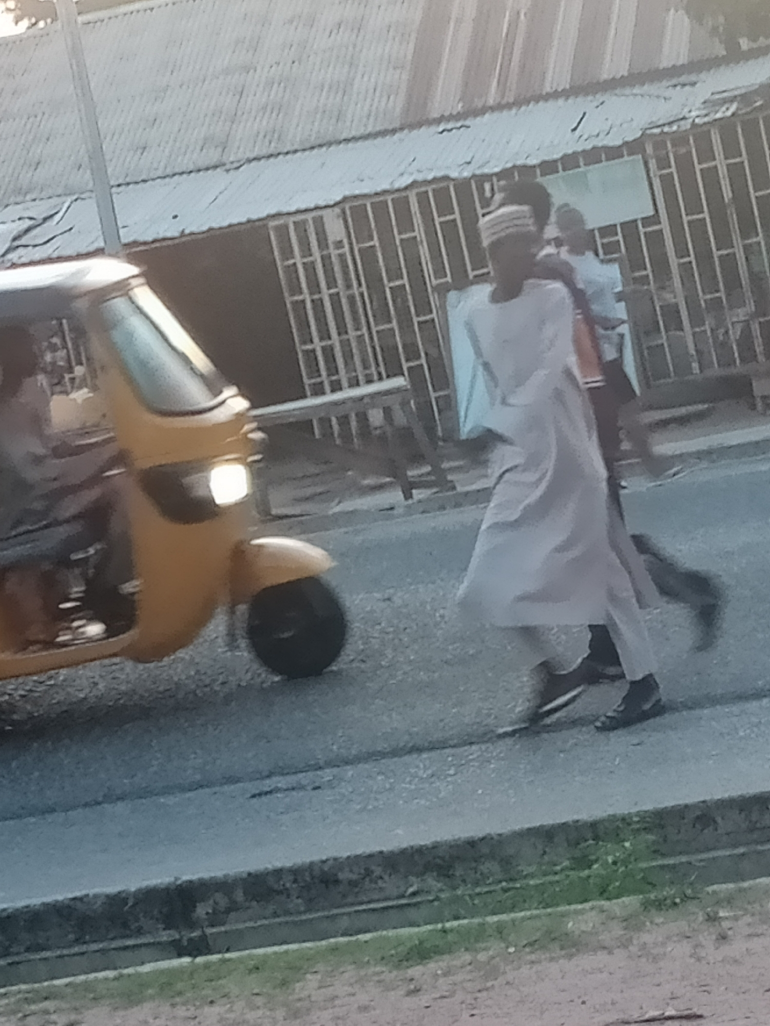 The image depicts a street scene in Jalingo, Nigeria. A yellow three-wheeled auto-rickshaw is stationary on the right side of the frame, with its headlight illuminated. A person is seated in the driver's position, partially visible. In the background, two individuals are walking. One person, wearing a light-colored garment and a striped head covering, is in the foreground, walking towards the right and slightly away from the camera. Another individual, wearing a light-colored shirt, is walking behind and to the right of the first person. The environment appears to be an urban setting with corrugated metal roofing and barred windows visible on structures in the background. The road surface is asphalt.