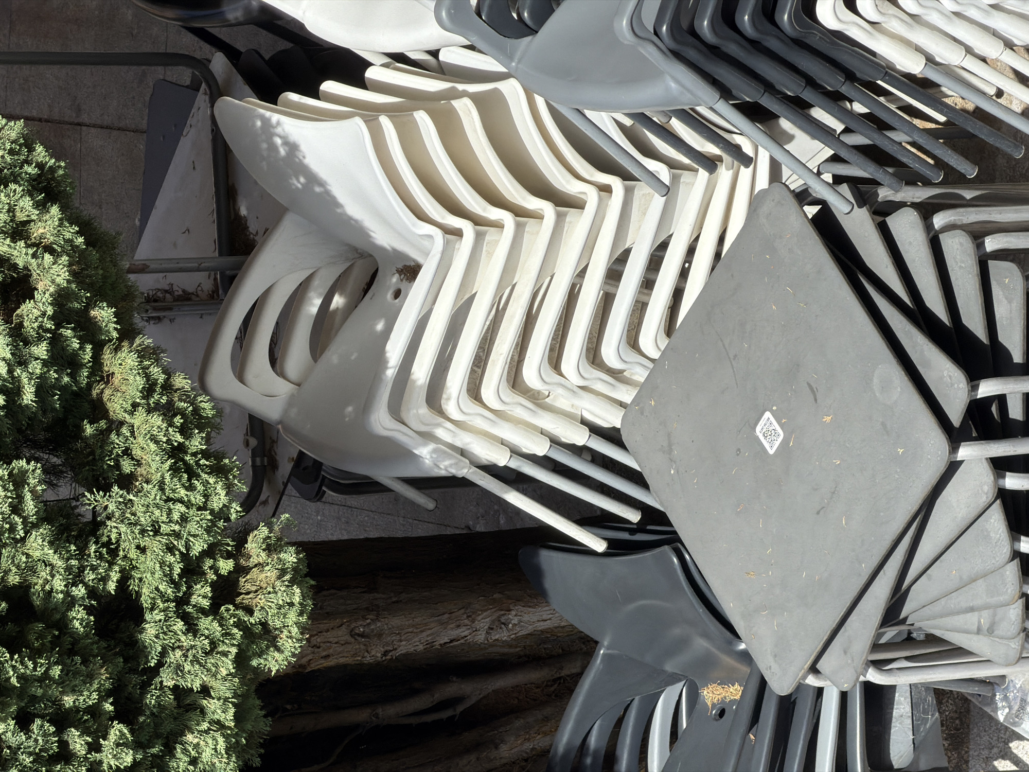 This image depicts a pile of stacked outdoor chairs and tables, likely in a storage area. The furniture is primarily made of plastic and metal. There are several stacks of white plastic chairs, a stack of dark grey plastic chairs, and a stack of dark grey square tables in the foreground. AQR code is visible on the top table. A large, mature evergreen tree with dark bark is situated to the left of the stacks. The ground appears to be paved with stone tiles. The scene is illuminated by natural sunlight, casting shadows on the furniture. The environment suggests an outdoor setting in a city, possibly a restaurant patio or a public space, in Las Rozas de Madrid, Spain. No people are present in the image.