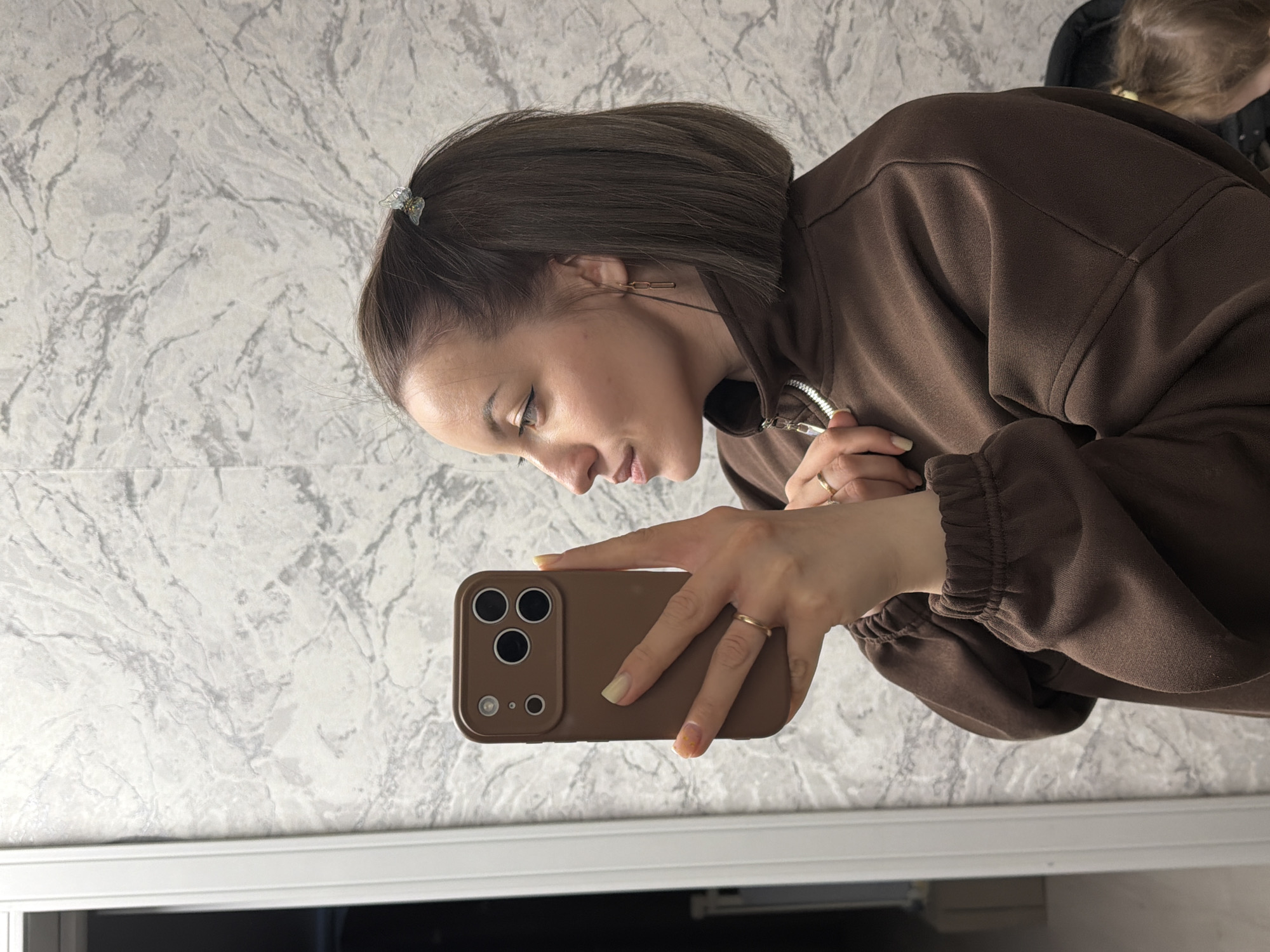 A woman with short brown hair, wearing a brown sweatshirt and gold jewelry, is holding a brown smartphone in her hands. She is looking at the screen of the phone. The background is a patterned wall. The location is likely indoors. Reshetnikovo, Russia, cannot be confirmed from the image.
