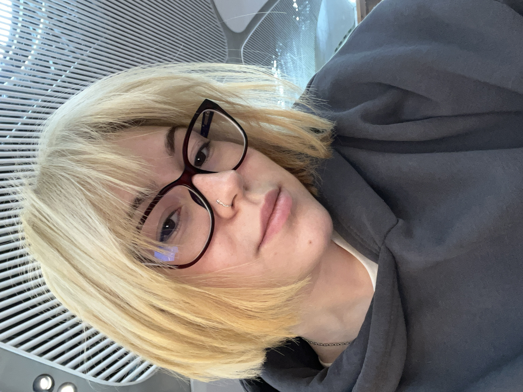 A blonde-haired individual with bangs and bob-length hair is featured in a close-up, tilted portrait. They are wearing dark-rimmed, subtly angular glasses and a small, decorative silver nose ring. The person looks directly at the camera with a neutral expression. They are dressed in a dark grey hooded sweatshirt, over what appears to be a lighter top, and a thin silver chain is visible around their neck.
The background reveals a modern indoor setting with distinctive white slatted or perforated panels on the ceiling or upper walls, creating a linear, grid-like pattern. Bright, even lighting illuminates the scene, with some small, circular lights visible in the lower left corner. The architectural style suggests a contemporary public building.
Given the location context of Arnavutköy, Turkey, this setting strongly points to Istanbul Airport, known for its modern design and prominent slatted structures. The image captures a moment of calm, with no specific activity other than posing for the photo. No visible text is present.