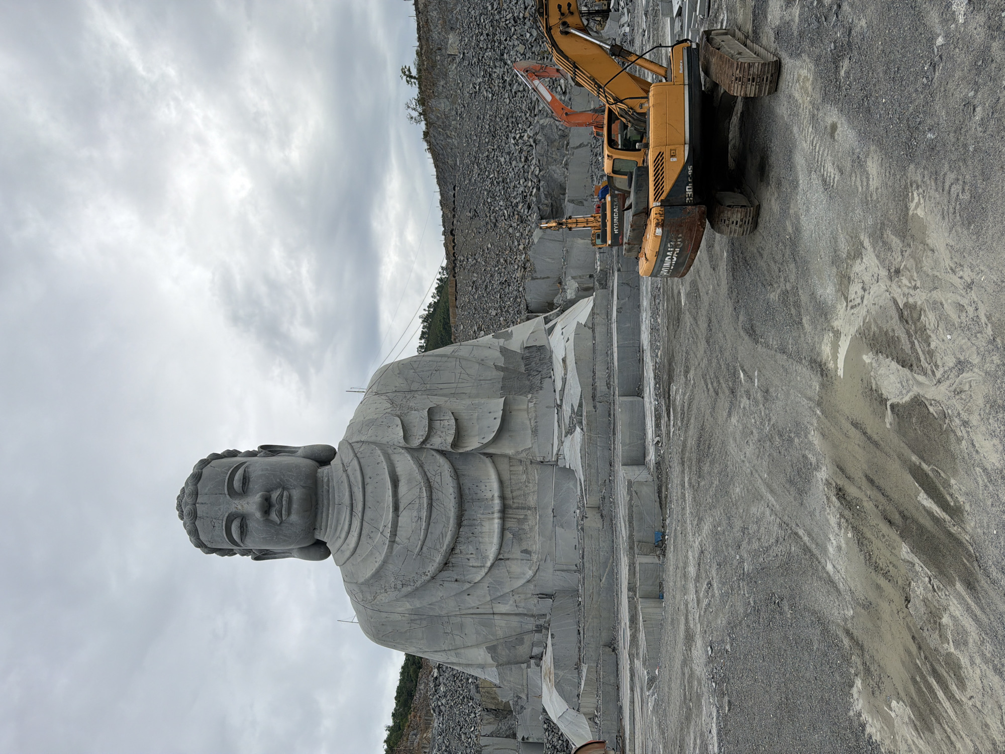 The image shows a large, unfinished stone Buddha statue in a quarry-like environment. The statue is gray and depicts the Buddha in a seated position. The quarry has machinery and excavated stone. The sky is cloudy. The location is most likely at the foot of the Marble Mountains on the outskirts of Đà Nẵng, Vietnam. There is no one shown in the picture. The picture may have been taken from ground level. The machinery is positioned near the statue.