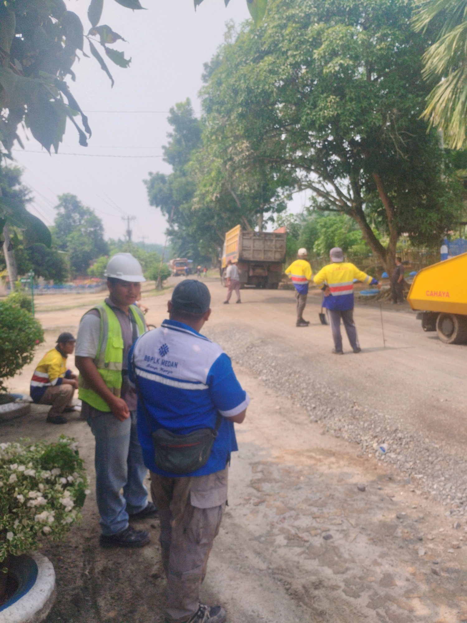 In Pagarbosi, Indonesia, a group of men, likely construction workers, are gathered near a road. Two men in the foreground are conversing. One man, wearing a white hard hat, a reflective yellow vest over a grey shirt, and blue jeans, stands facing away from the camera. The second man, from the back, wears a blue and white polo shirt with the text "BBPLK MEDAN" and has a dark fanny pack around his waist. He is also wearing khaki pants and dark shoes. To the left, a third man in a yellow and blue striped jacket and a baseball cap is seated. In the background, three men in similar yellow and blue jackets are standing near a yellow dump truck. Another yellow construction vehicle, possibly a compactor, is visible on the right, with the word "CAHAYA" written on its side. The environment appears to be a rural or semi-rural area with a gravel road, trees, and vegetation. The sky is overcast.