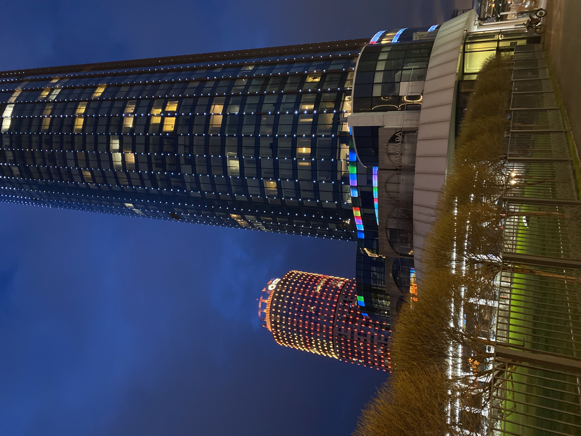 The image presents a striking night view of two illuminated, modern high-rise buildings in Yekaterinburg, Russia, against a deep blue sky. The photograph is oriented vertically but depicts the buildings as if they are standing upright, showcasing their architectural brilliance.
The prominent building on the left is a tall, curved glass skyscraper, likely the Hyatt Regency Ekaterinburg. Its facade is distinguished by a grid of numerous small, white and blue lights that create a shimmering effect. Several windows glow with warm yellow light from within. Towards its base, colorful vertical stripes of red, green, and blue lights are visible, along with a sign that clearly displays the text "УРАЛ" (URAL). Reflections of the night sky and surrounding lights are visible on its glass surfaces.
Behind and to the right of this main structure stands a second, shorter, cylindrical building. It is entirely covered in a vibrant, intricate pattern of red and yellow lights, with a large, illuminated letter, possibly 'G', near its top. This is identifiable as part of the Yekaterinburg-EXPO complex.
In the foreground, a low, light brown hedge or row of sparse trees runs horizontally, separating the viewer from the scene. Behind it, a metal fence and a patch of green grass are visible. The overall scene captures a moment of urban tranquility, highlighting the modern, illuminated architecture of the city at night. No people or specific activities are discernible.