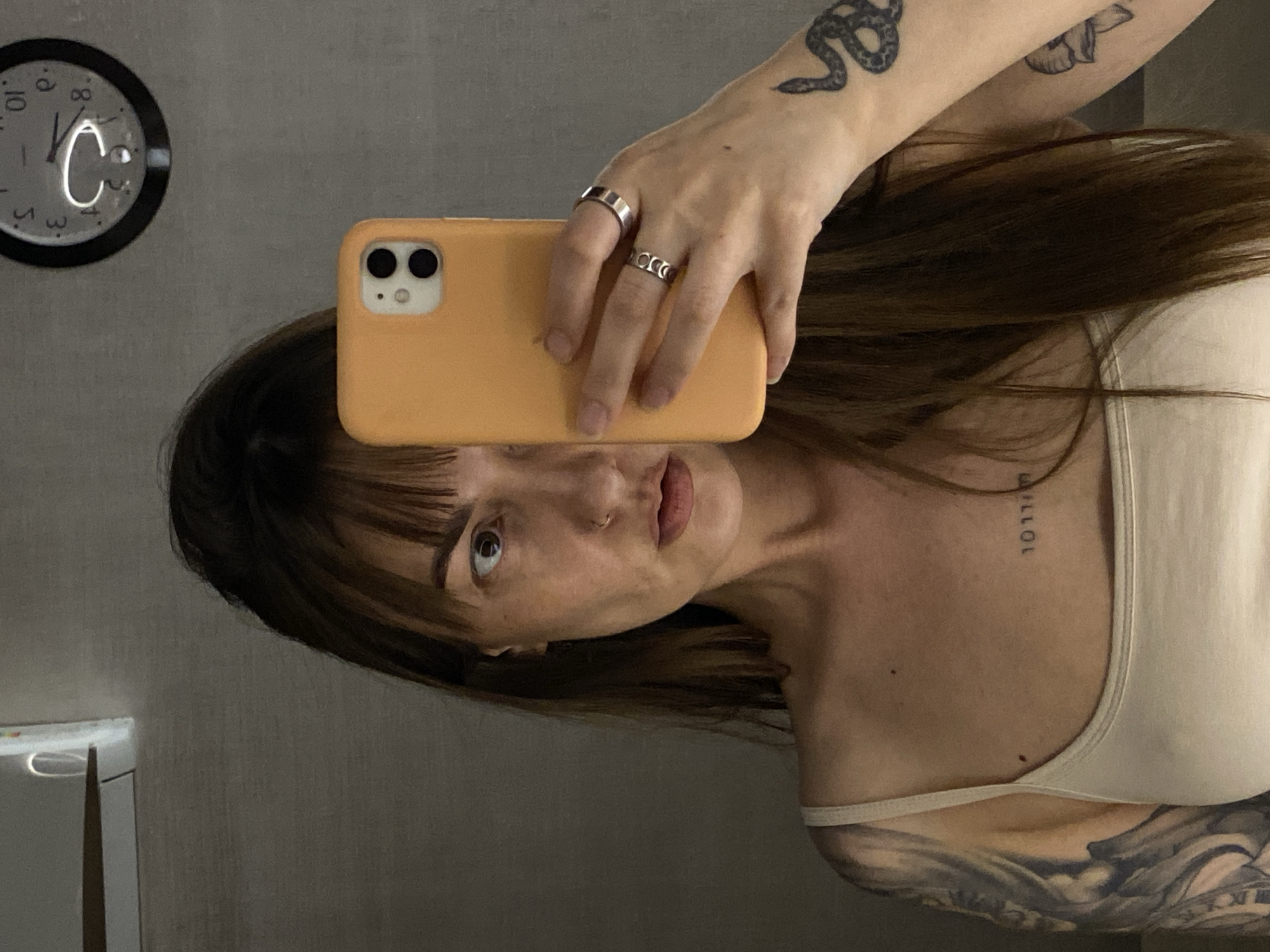 A woman is taking a selfie in front of a mirror. The phone is orange and obscures part of her face. She has dark hair with bangs, light skin, and multiple tattoos, including one of a snake on her left arm and writing on her chest. She's wearing a light-colored tank top. On the wall behind her is a clock. The environment appears to be a bathroom with gray walls. There is no information to suggest that this image was taken in Votkinsk, Russia.