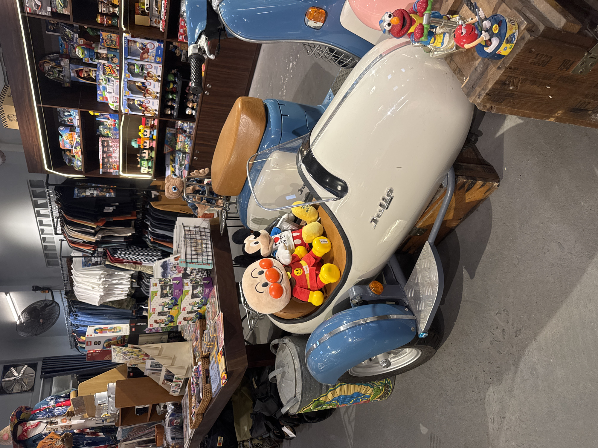 The image shows the interior of a store in Nha Trang, Vietnam. The store appears to be a novelty or toy shop, with shelves stocked with various toys and collectibles, including figures from popular media like Toy Story and possibly characters from children's shows. In the foreground, a white three-wheeled vehicle resembling a miniature motorcycle with a sidecar is prominently displayed. Two plush toys are seated in the sidecar: one is Anpanman, a popular Japanese cartoon character, and the other is Mickey Mouse. Behind the vehicle, a blue scooter is parked. The environment is well-lit with artificial lighting, and racks of clothing are visible in the background, suggesting the store may also sell apparel. The floor is concrete, and wooden crates are used as display stands for some of the items.