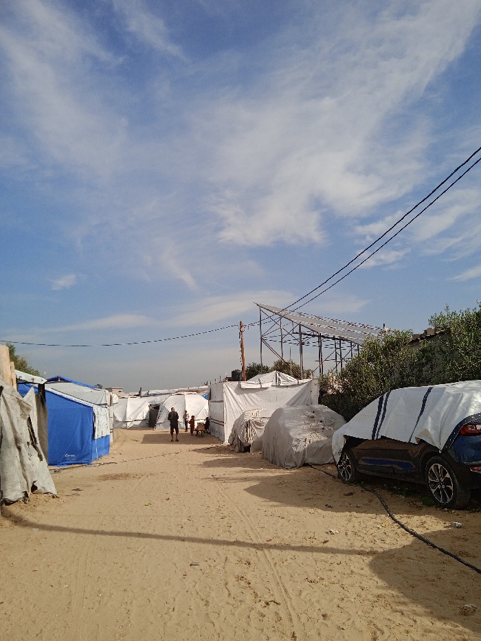 This image depicts an unpaved sandy path winding through a densely constructed makeshift camp in Khan Yunis, Palestinian Territory, under a clear blue sky with scattered white clouds. Numerous shelters, primarily constructed from white tarpaulins and some blue fabric, line both sides of the path. In the mid-ground, an adult male wearing a dark shirt and shorts stands facing away from the camera, near a small group of children gathered beside a shelter. To the right, two vehicles are partially covered with white tarps; the closest is a dark blue SUV with visible silver wheels, from which a black cable or hose extends onto the sand. Further right, green foliage is visible behind a concrete wall. In the background on the right, a large metal framework supports multiple dark panels, possibly solar panels, mounted on a raised structure. Utility poles with overhead wires traverse the scene.